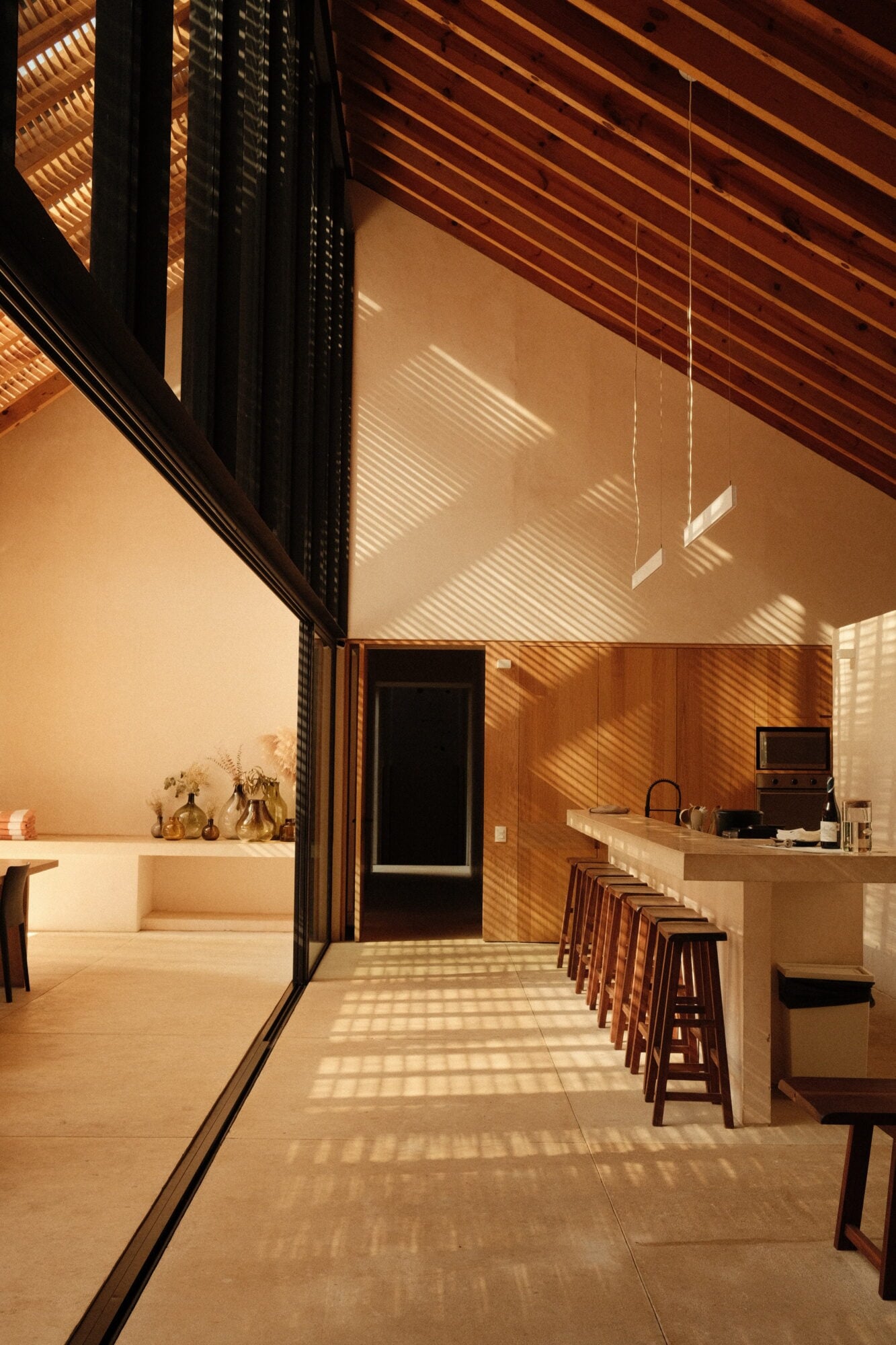Casa Zarzales, Mexico — Minimalist kitchen with wooden beams, exposed ceiling, concrete counters, bar seating, and geometric sunlight patterns across cream walls.