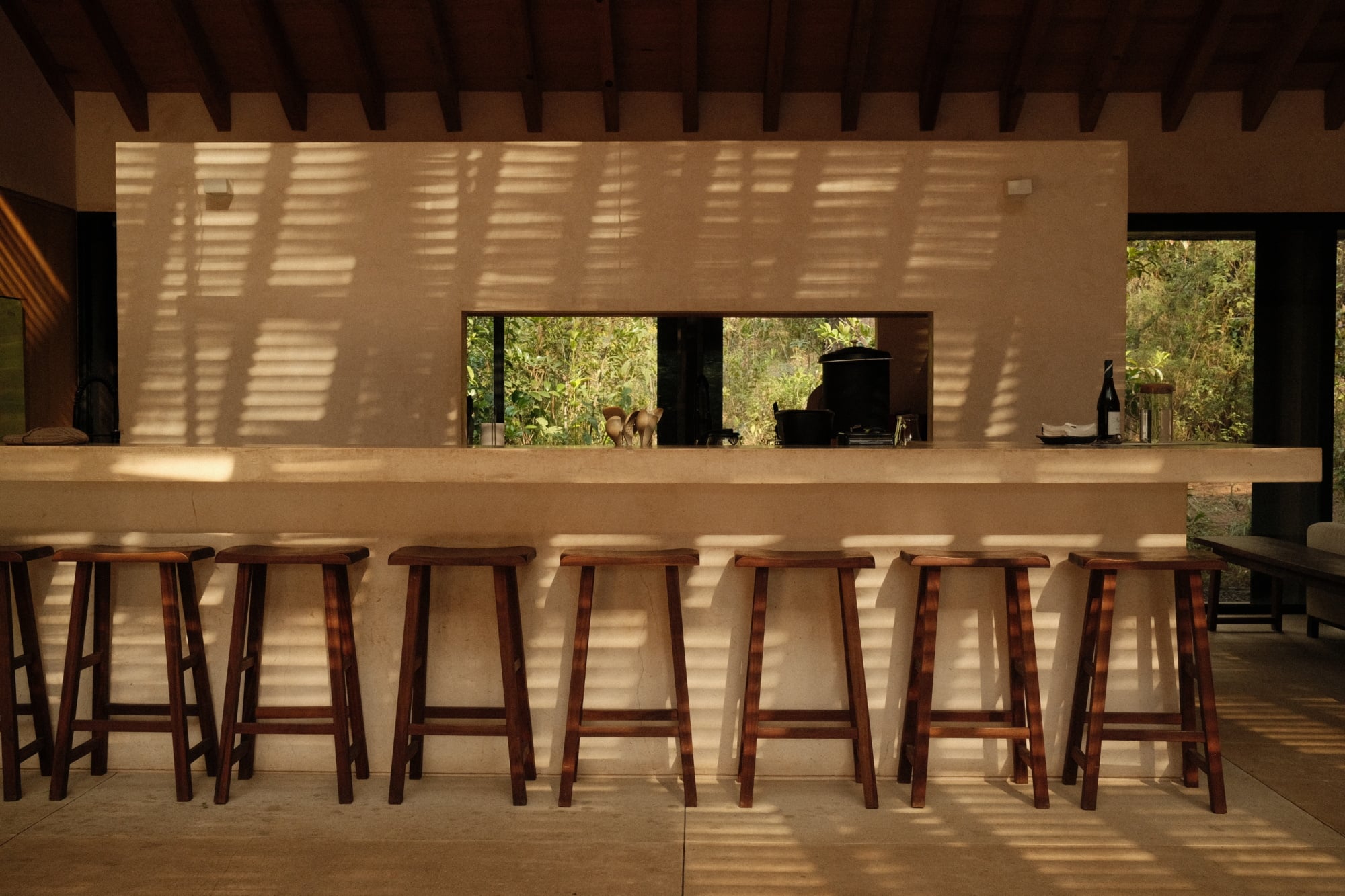 Casa Zarzales, Mexico — Minimalist concrete bar with wooden stools, louvered ceiling casting linear shadows, open kitchen visible beyond, lush landscape framed through windows.