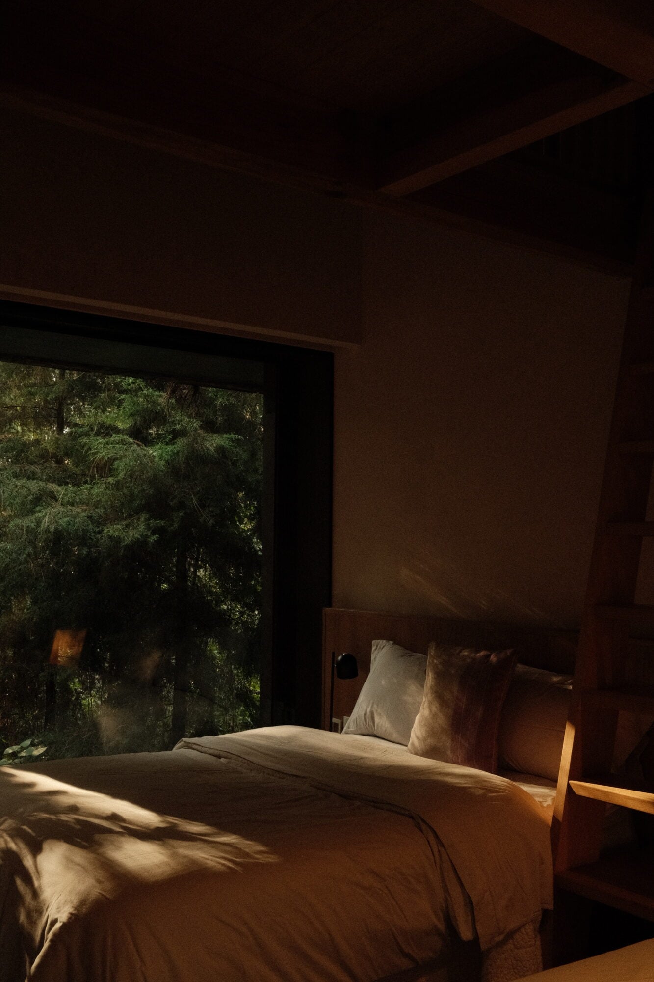 Casa Zarzales, Mexico — Minimalist bedroom with wooden bed frame, terracotta walls, floor-to-ceiling window framing dense forest canopy, warm afternoon light casting shadows across neutral linens.