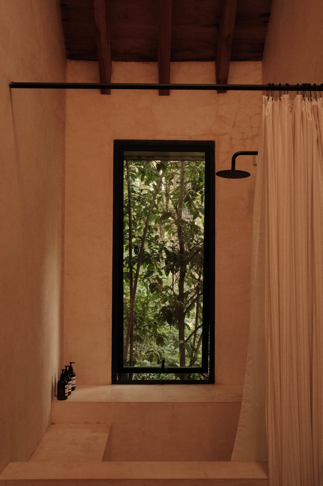 Casa Zarzales, Mexico — Minimal bathroom with terracotta walls, black-framed window opening onto lush forest, rainfall showerhead, cream linen curtain.