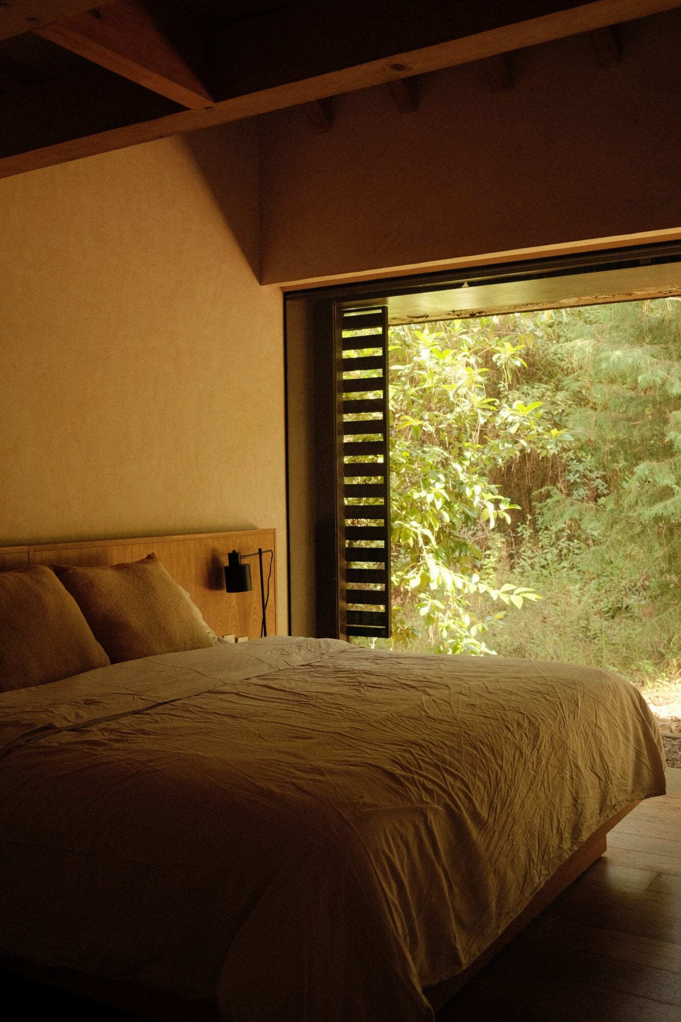 Casa Zarzales, Mexico — Minimalist bedroom with warm ochre walls, wooden ceiling beams, and floor-to-ceiling slatted window overlooking lush forest canopy.