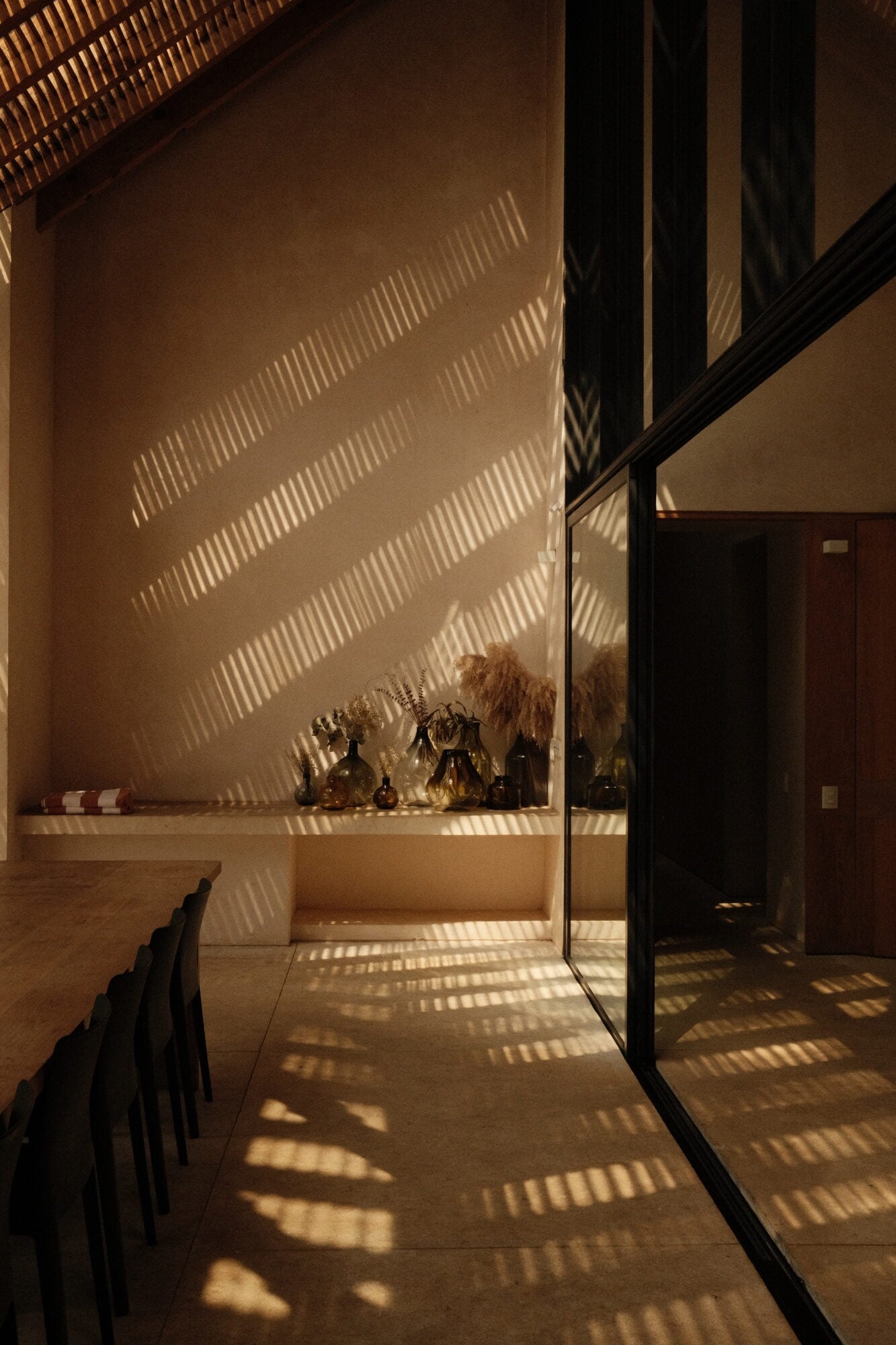 Casa Zarzales, Mexico — Minimalist dining space with slatted light filtering across cream walls, black-framed glass doors, and sculptural vessels on floating shelf.