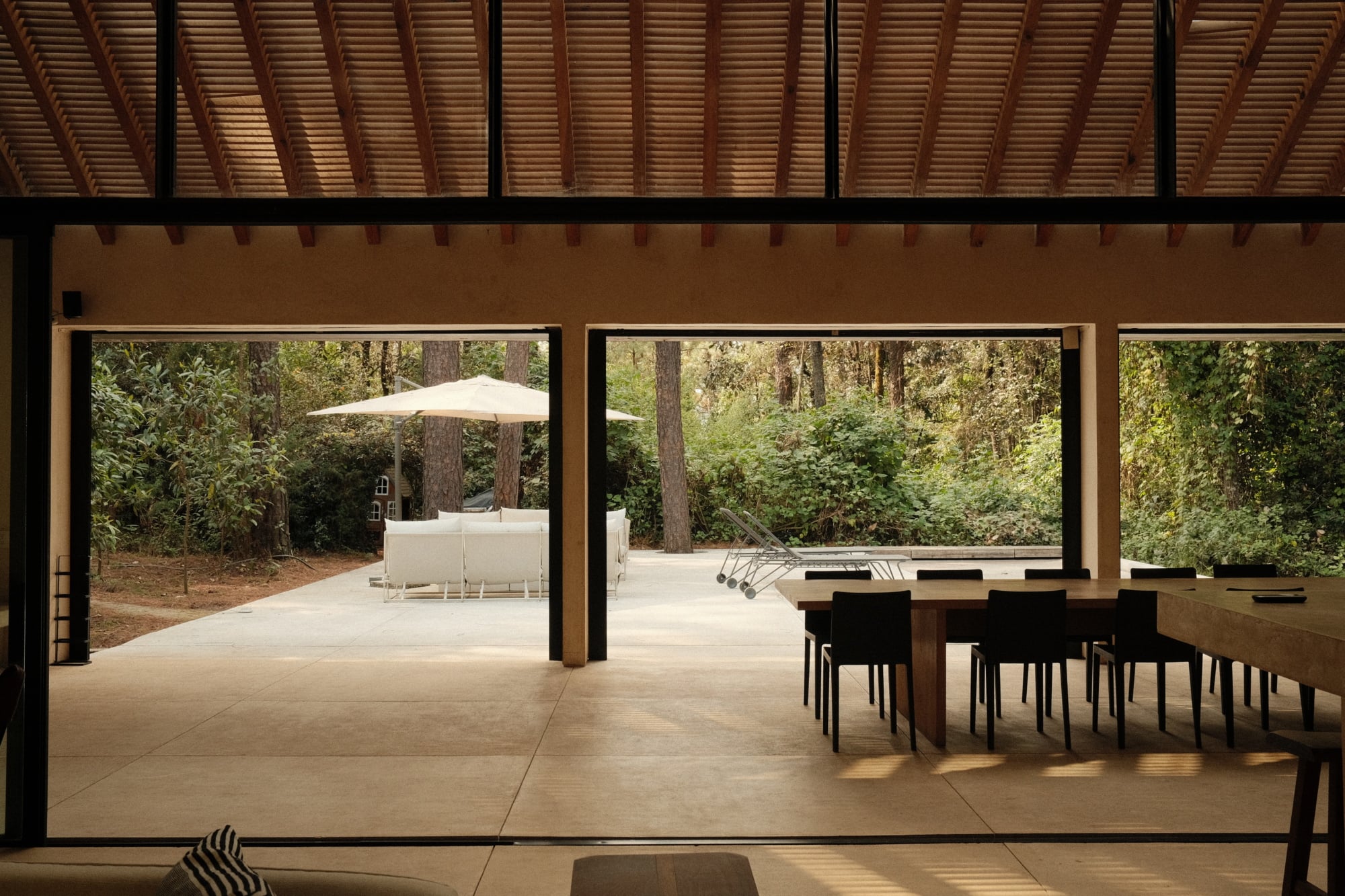 Casa Zarzales, Mexico — Open-plan dining pavilion with timber ceiling, concrete floors, and floor-to-ceiling glass doors opening to forest landscape and terrace with umbrella and lounge chairs.