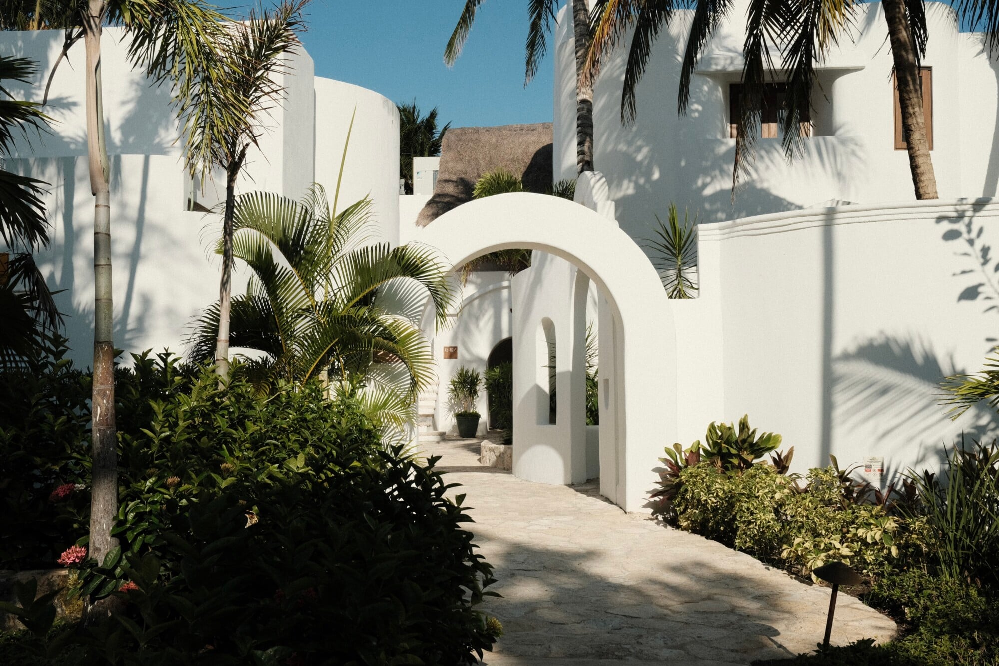 Maroma, A Belmond Hotel, Mexico — White-walled courtyard with curved archway, palm trees casting shadows, tropical plants, minimalist architecture and bright natural light.