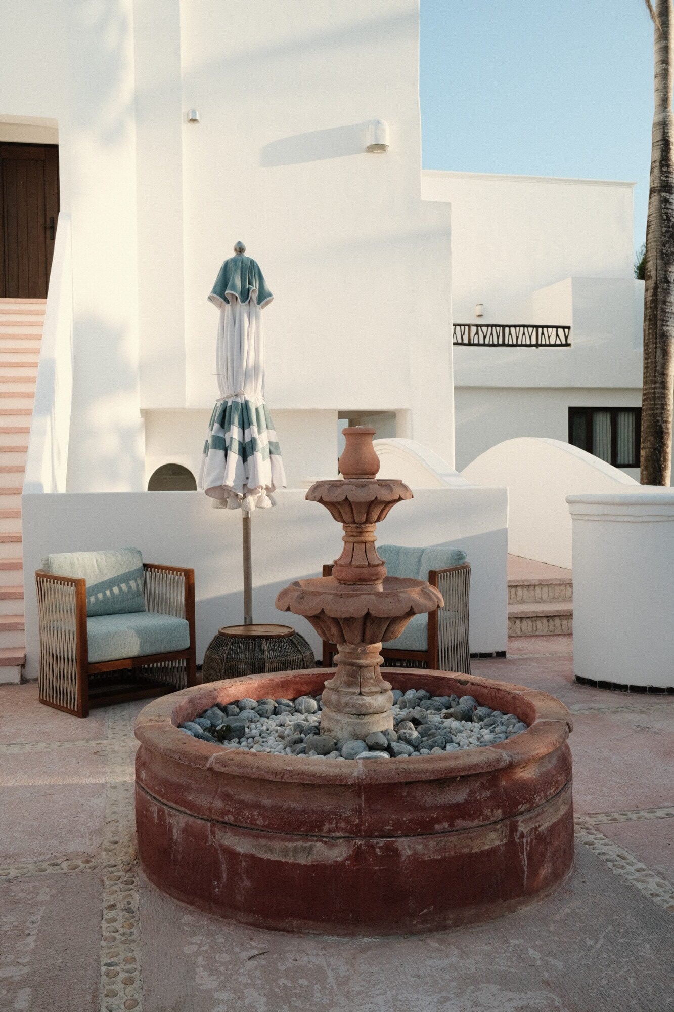 Maroma, A Belmond Hotel, Mexico — Whitewashed courtyard with terracotta fountain, striped umbrella, woven chairs, and clear blue sky casting geometric shadows.