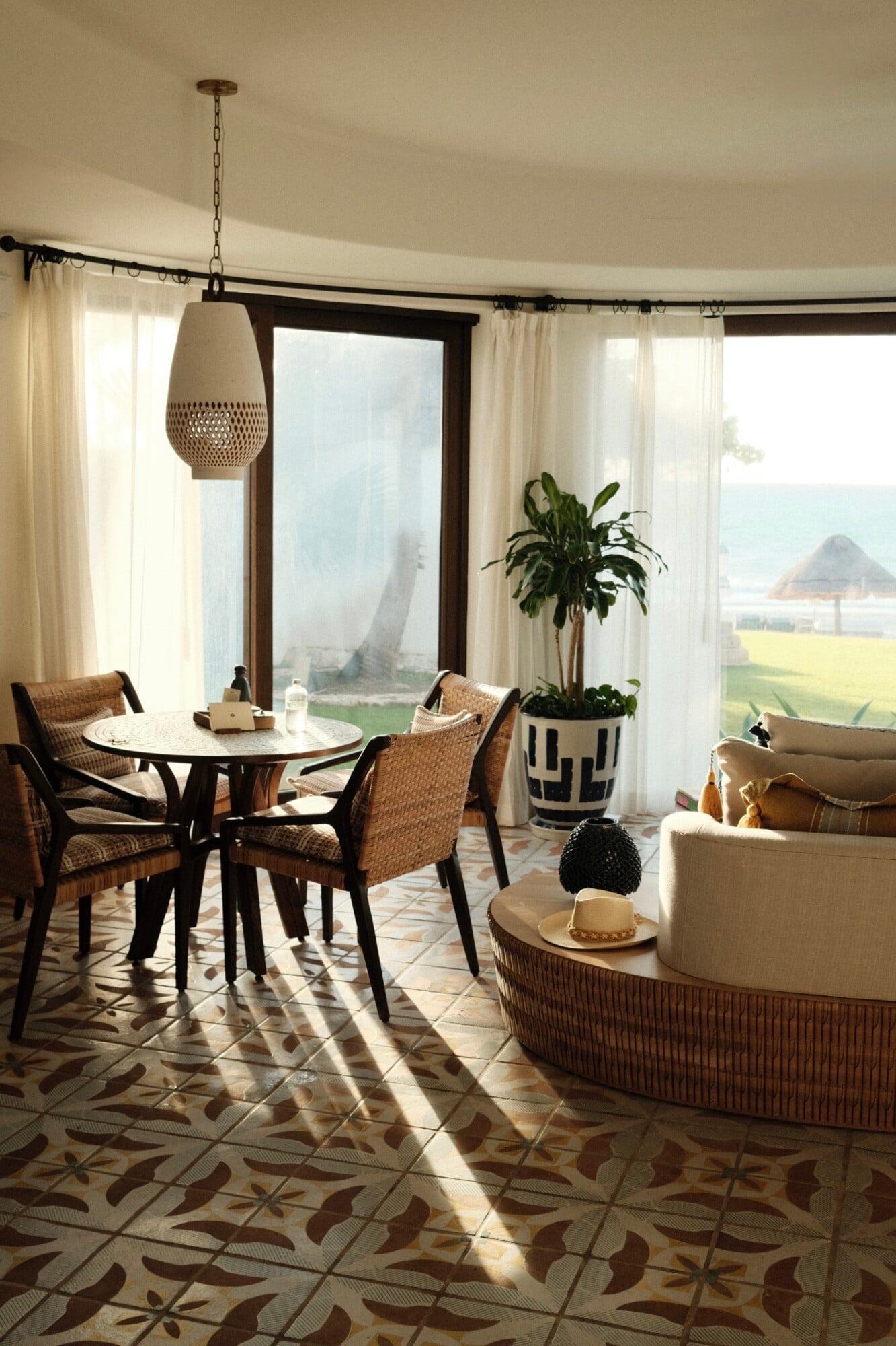 Maroma, A Belmond Hotel, Mexico — Curved glass doors frame ocean views in sunlit sitting room with woven chairs, patterned tile floor, cream curtains, and natural light casting geometric shadows.