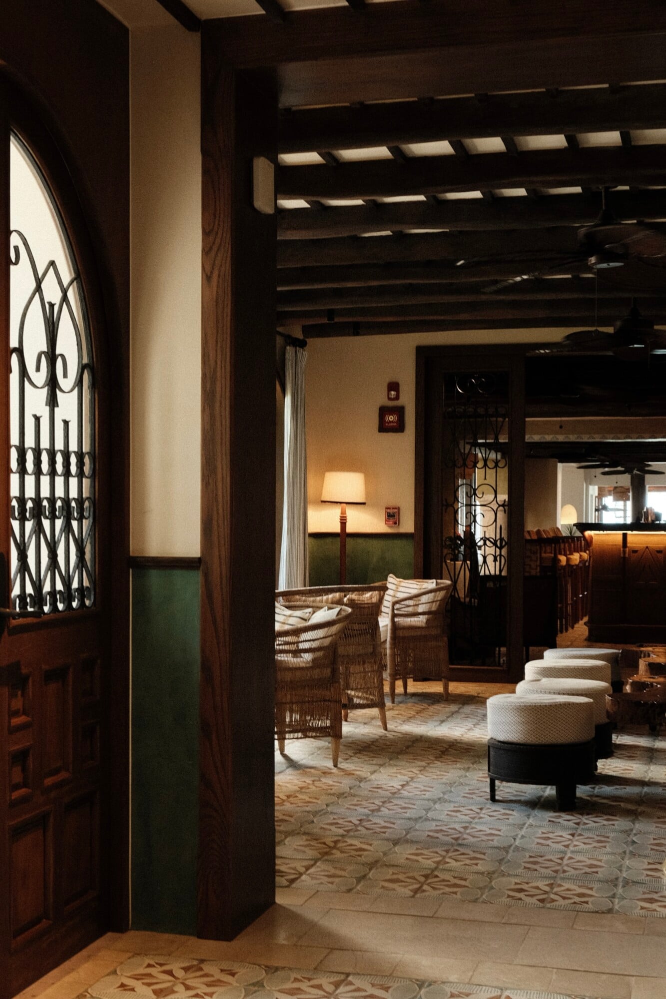 Bambuco by Handshake, Mexico — Dark wood beams and wrought-iron details frame a warm hospitality interior with woven seating, geometric tile flooring, and filtered natural light casting geometric shadows.