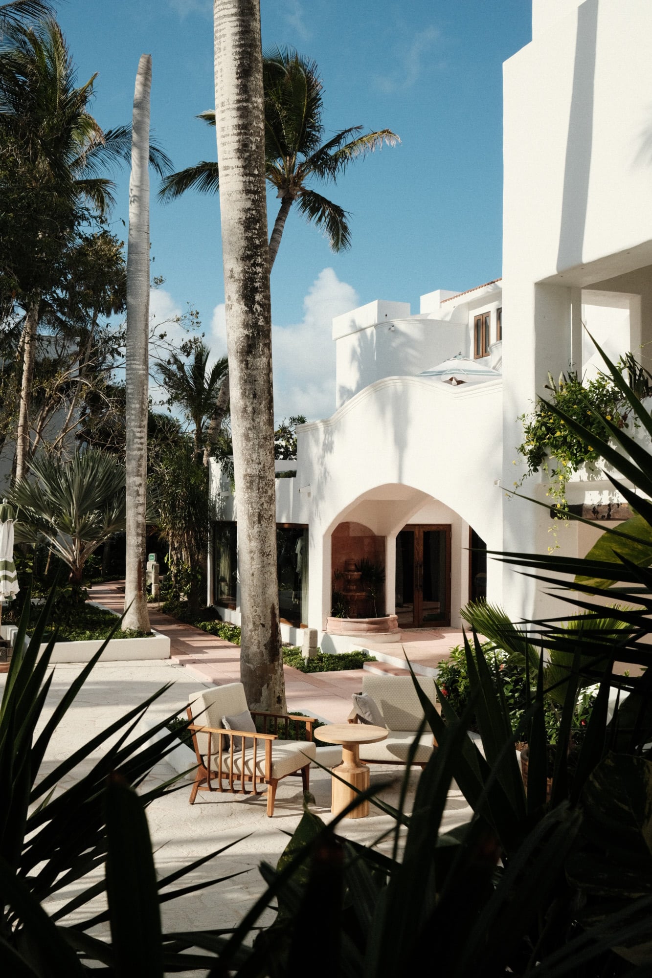 Maroma, A Belmond Hotel, Mexico — White-walled modernist courtyard with arched entryway, tall palm trees, terracotta floors, and mid-century wooden seating under clear blue sky.