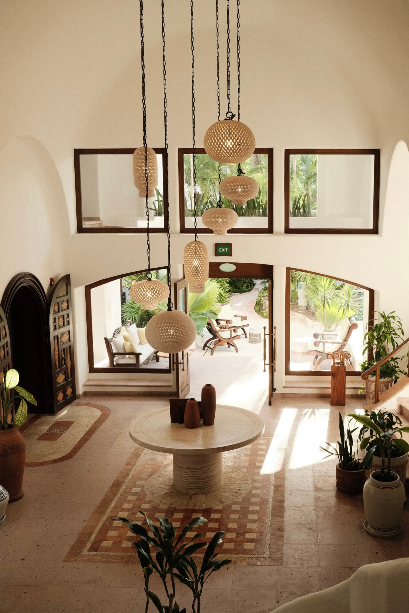 Maroma, A Belmond Hotel, Mexico — Woven pendant lights hang above a circular stone table in a bright coastal lobby with dark wood-framed mirrors, potted plants, and views to tropical gardens.