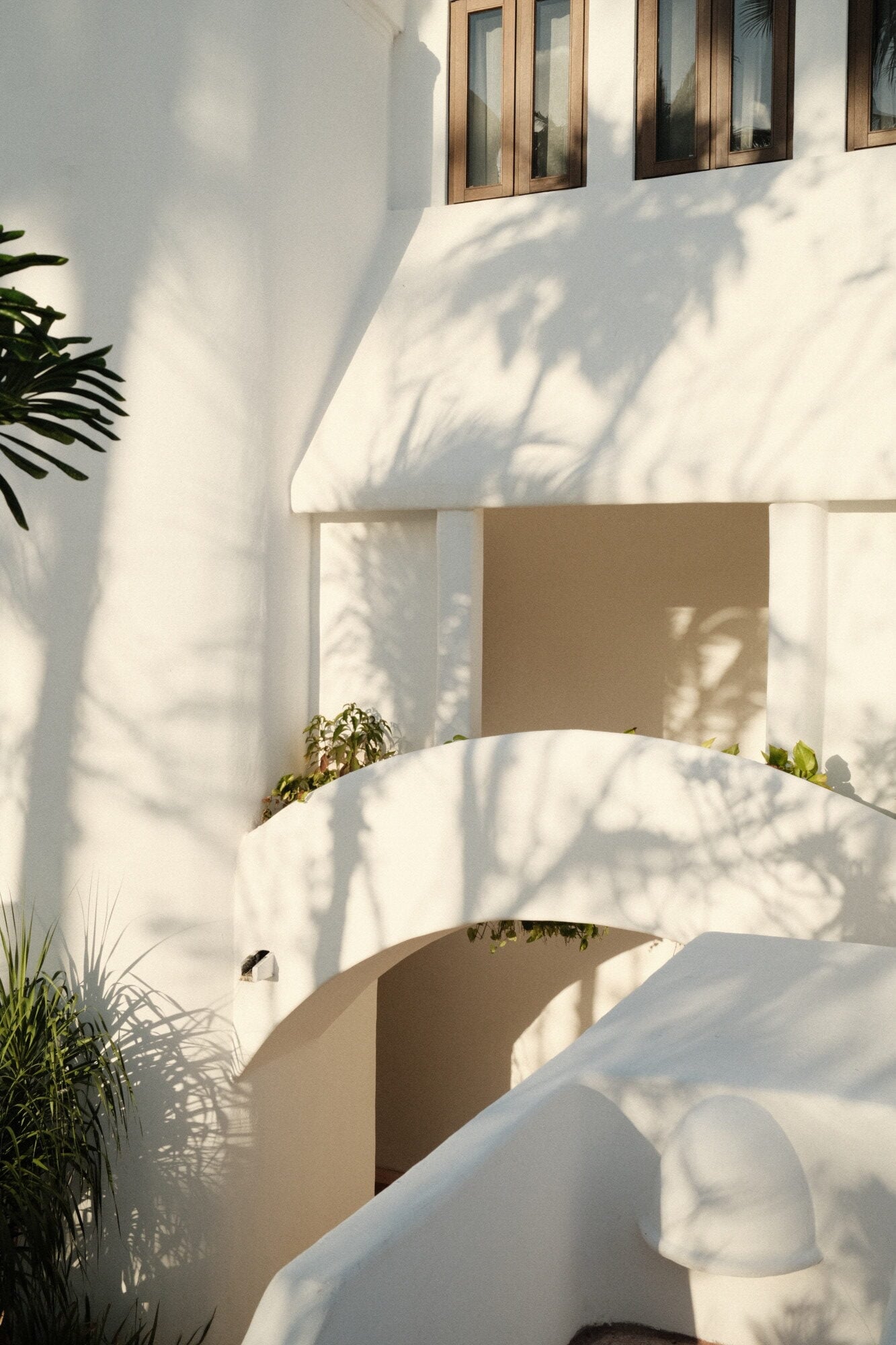 Maroma, A Belmond Hotel, Mexico — Minimalist white stucco architecture with curved walls, wooden-framed windows, dappled shadow patterns, and potted tropical plants in bright sunlight.