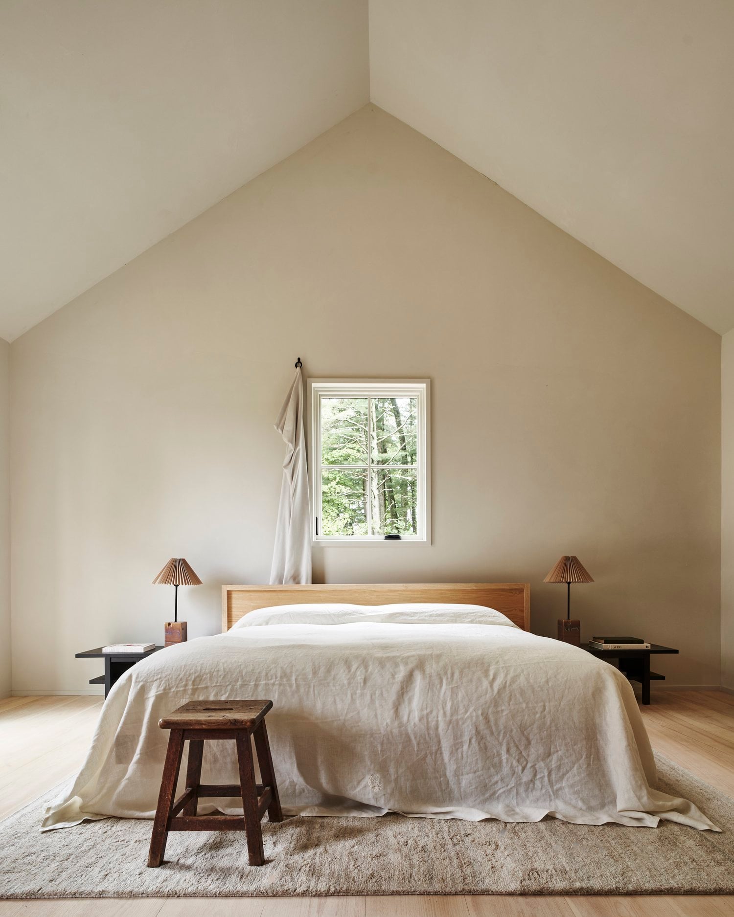 Inness, North America — Minimalist attic bedroom with natural wood bed frame, cream linens, pitched ceiling, and forest views through centered window.