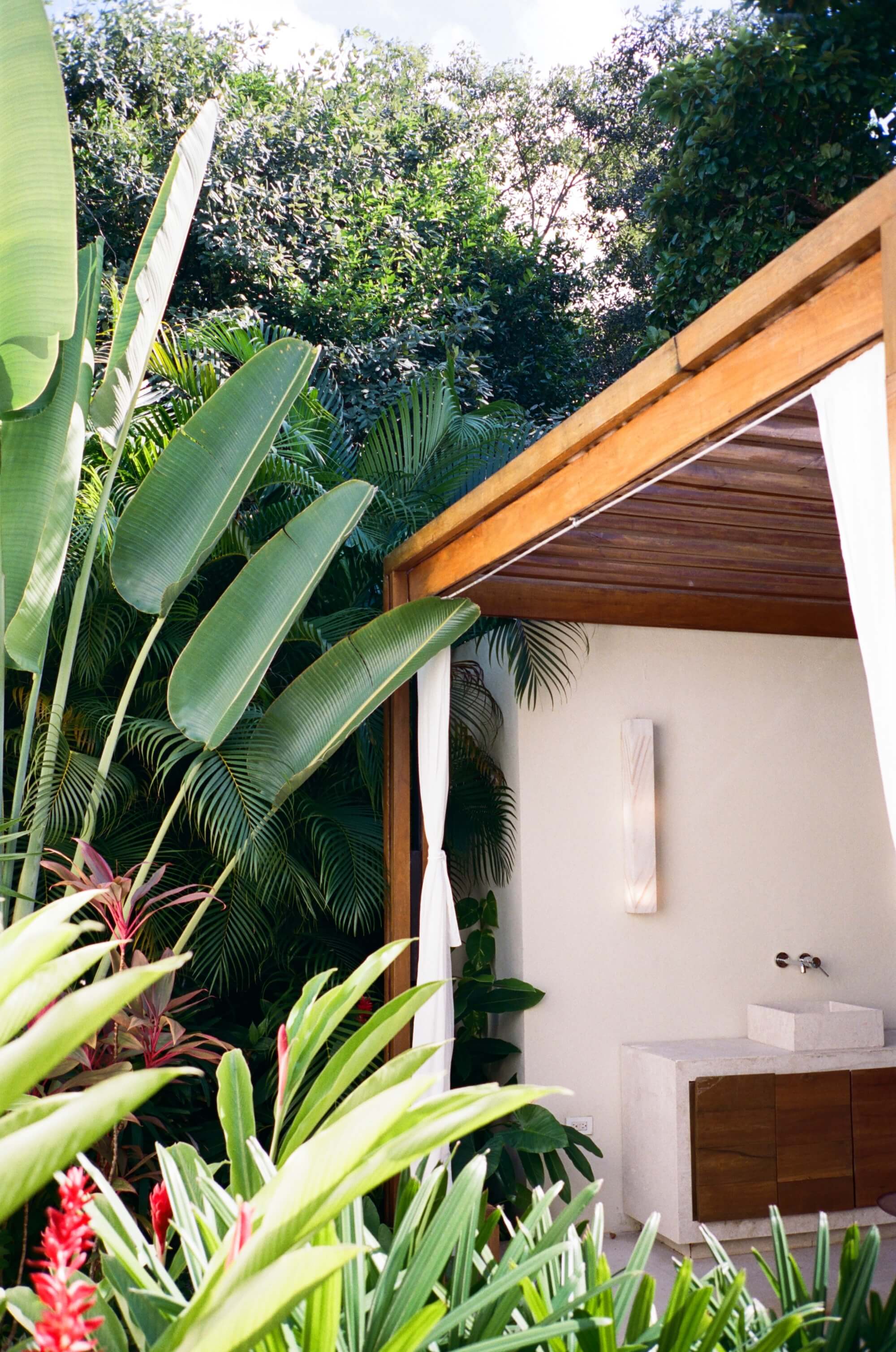 Rosewood Mayakoba, Mexico — Modern bathroom pavilion with teak ceiling, white walls, and lush tropical garden featuring banana plants and red ginger flowers.