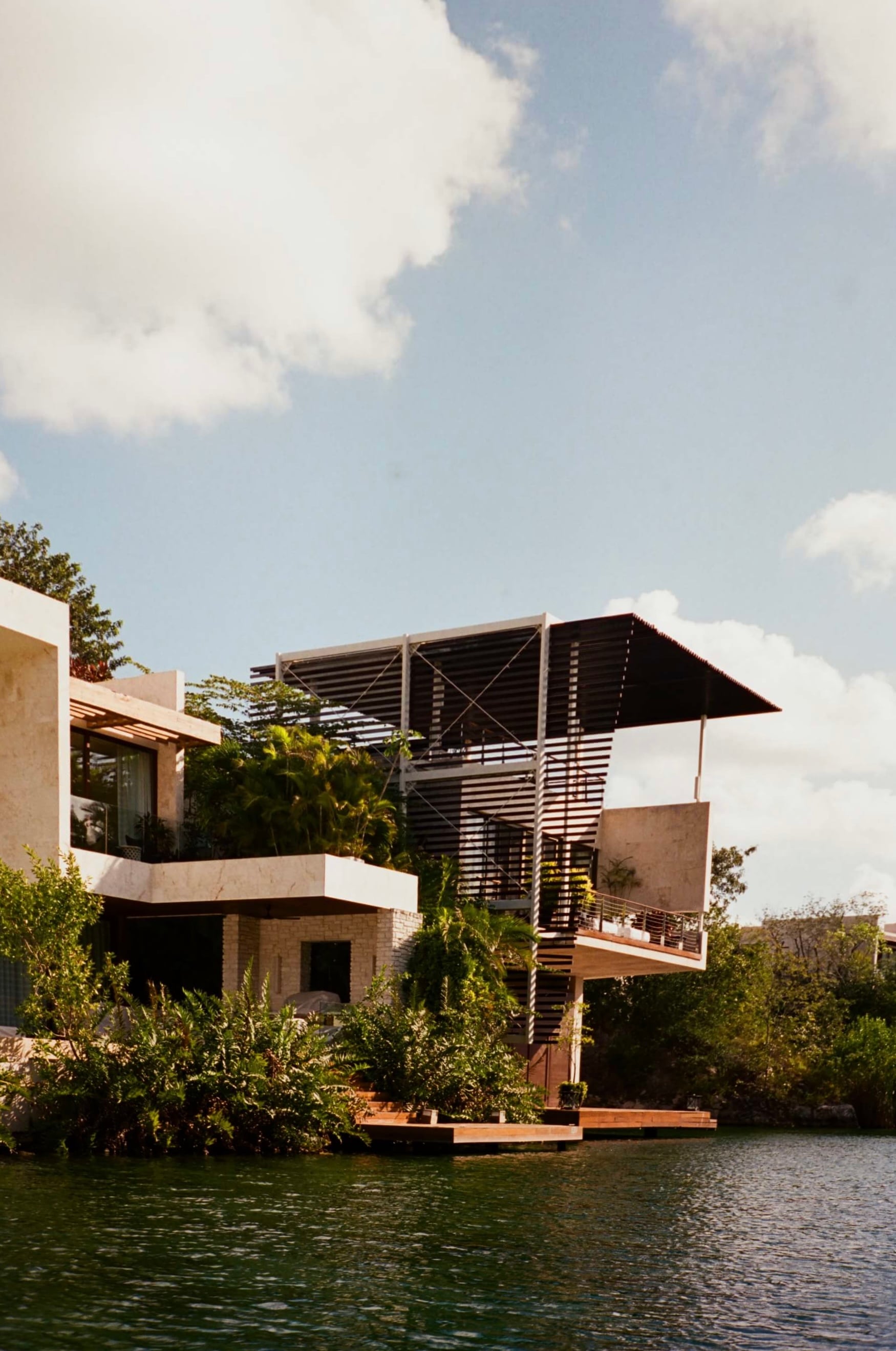 Rosewood Mayakoba, Mexico — Modernist waterfront residence with stacked terraces, dark timber screens, and earth-toned stone overlooking lagoon vegetation under blue sky.