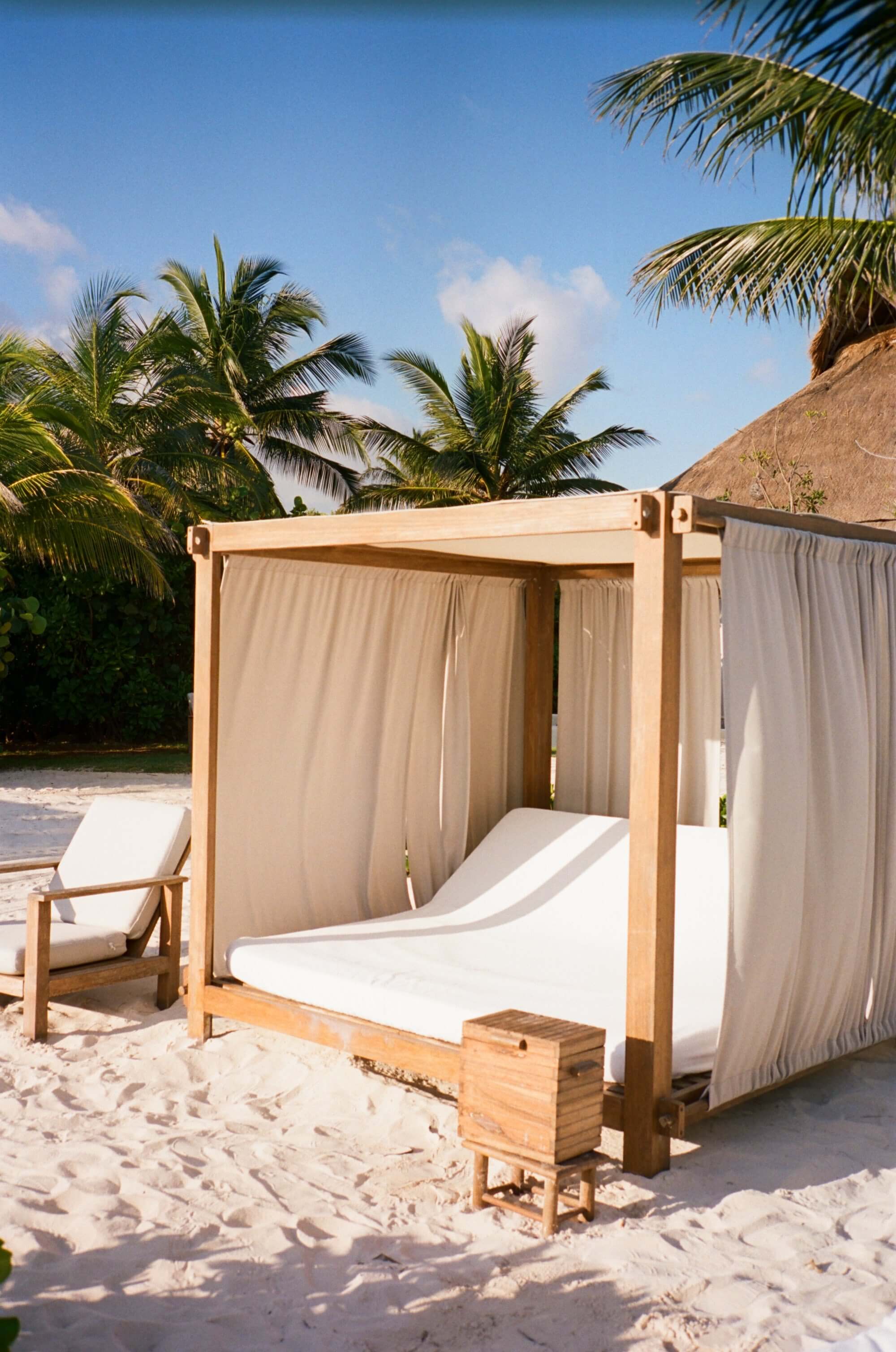 Rosewood Mayakoba, Mexico — Wooden canopy bed with flowing linen curtains on white sand beach with palm trees and thatched-roof structures.