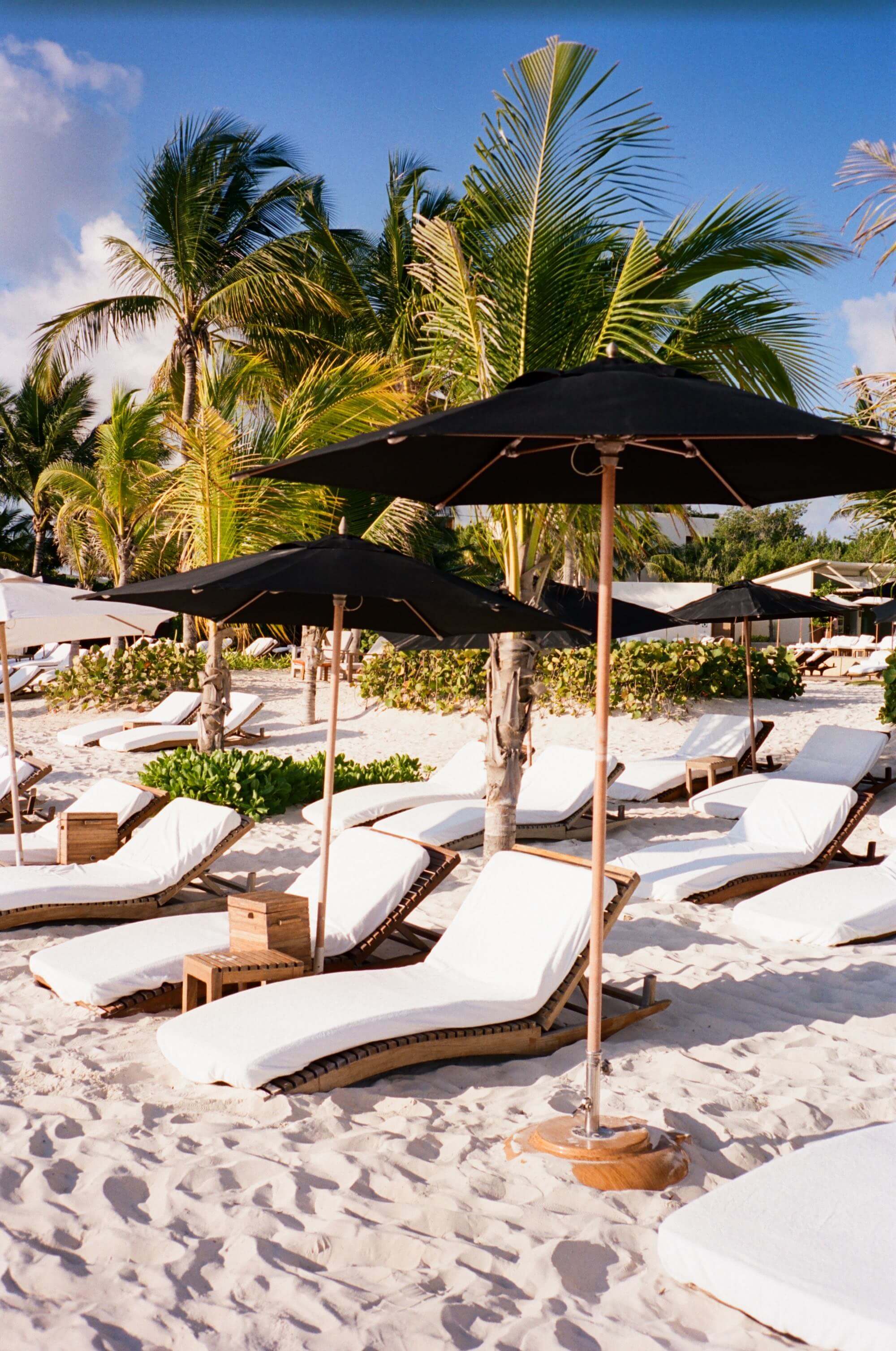 Rosewood Mayakoba, Mexico — Black umbrellas and teak loungers on white sand beach with palm trees and clear blue sky, luxury resort seating area.