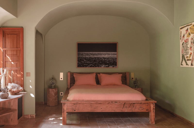 Hotel Corazón, Balearic Islands — Arched bedroom with terracotta wood bed, coral linens, sage walls, and natural light through shuttered door.