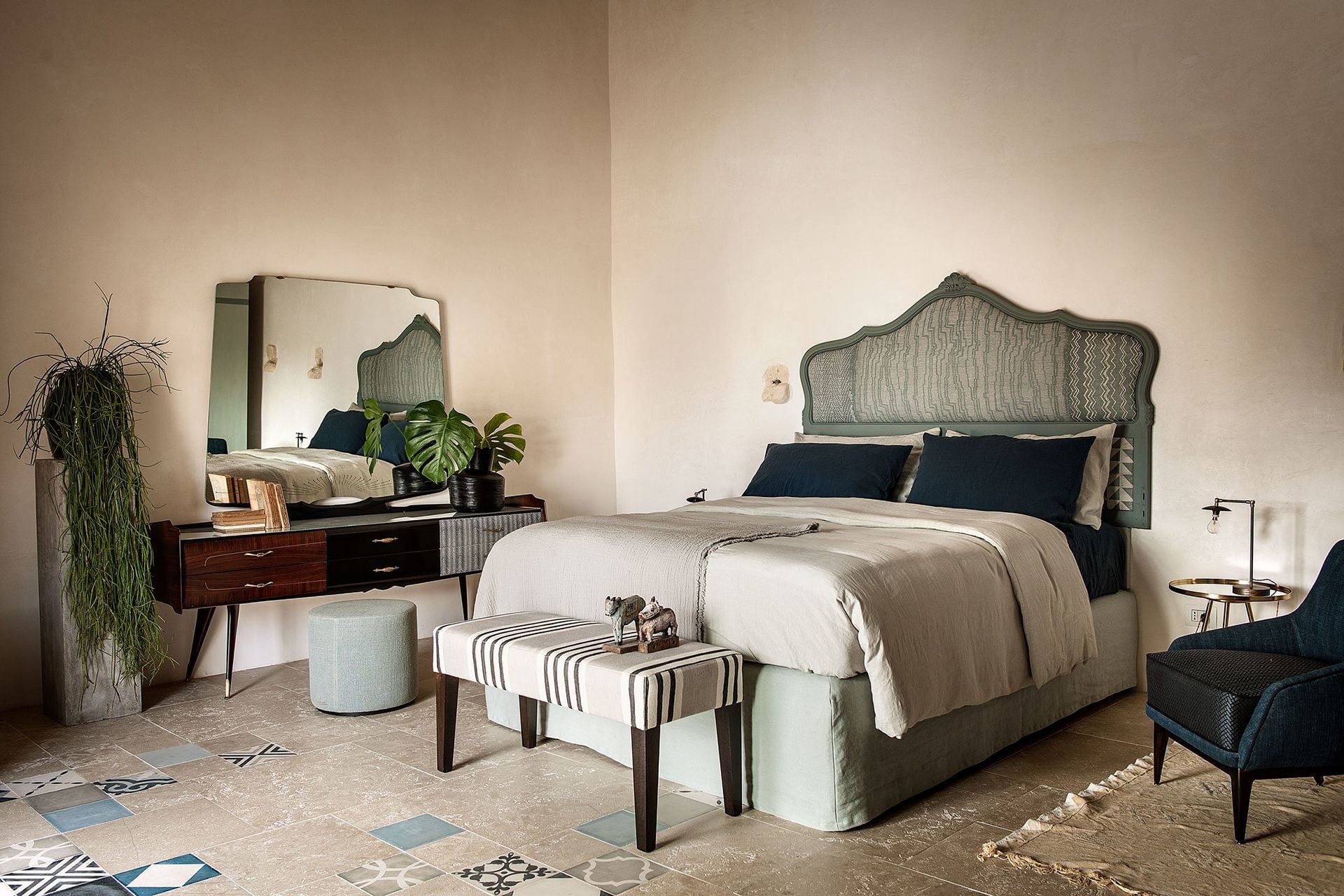 Dimora delle Balze, Europe — Modernist bedroom with green upholstered headboard, cream bedding, mid-century vanity, potted monstera, striped bench, and patterned terrazzo floors.