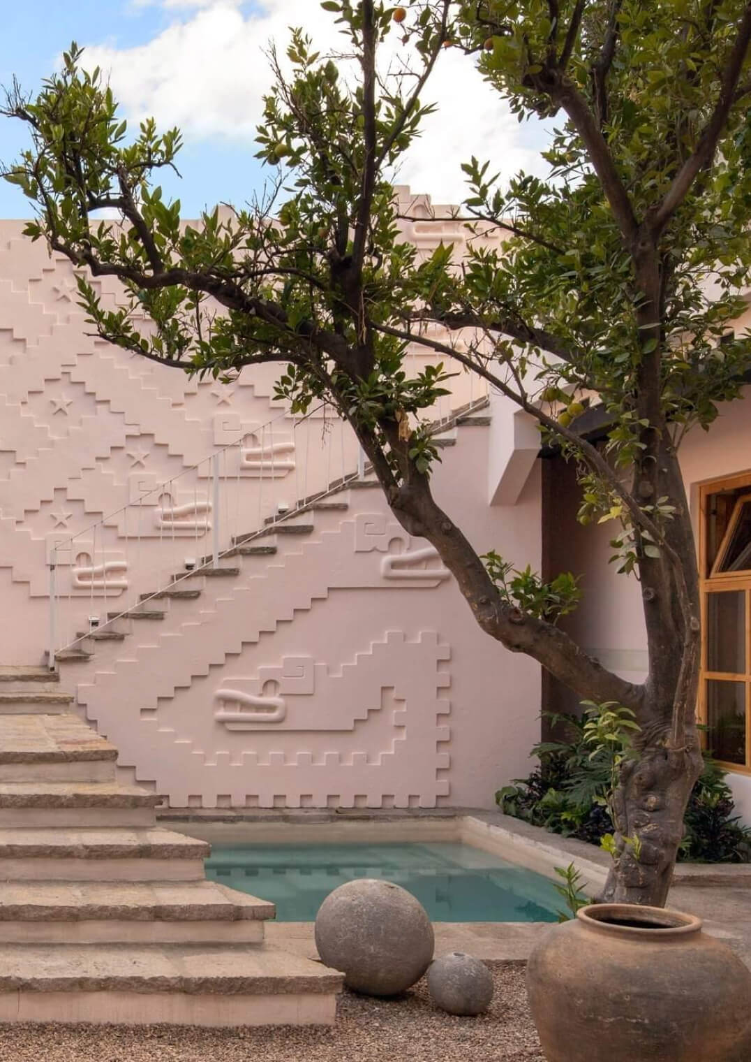 La Valise San Miguel, Mexico — Pink stucco courtyard with geometric relief wall, stone steps, mature tree, turquoise pool, and terracotta pottery accents.