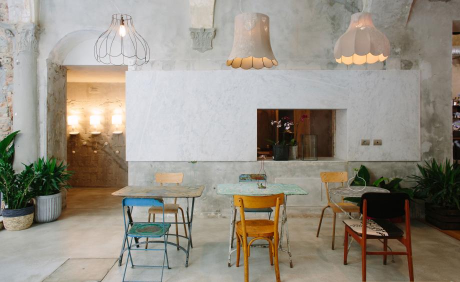 La Ménagère, Florence, Italy — Minimalist dining room with concrete walls, pendant lights, vintage wooden chairs, potted plants, and natural light from adjoining passage.