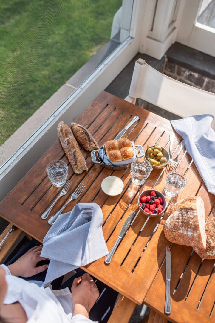 The Lake House, North America — Overhead view of wooden table set with fresh bread, berries, olives, and glassware beside open doors overlooking green lawn.