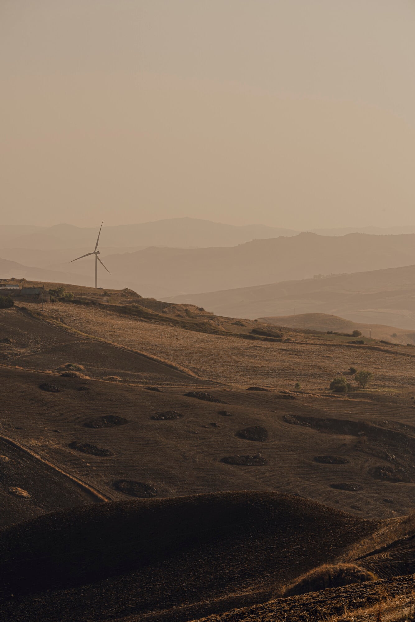 Susafa, Italy — Wind turbine on terraced hillside at golden hour, layered mountains in haze, agricultural landscape with dramatic light.