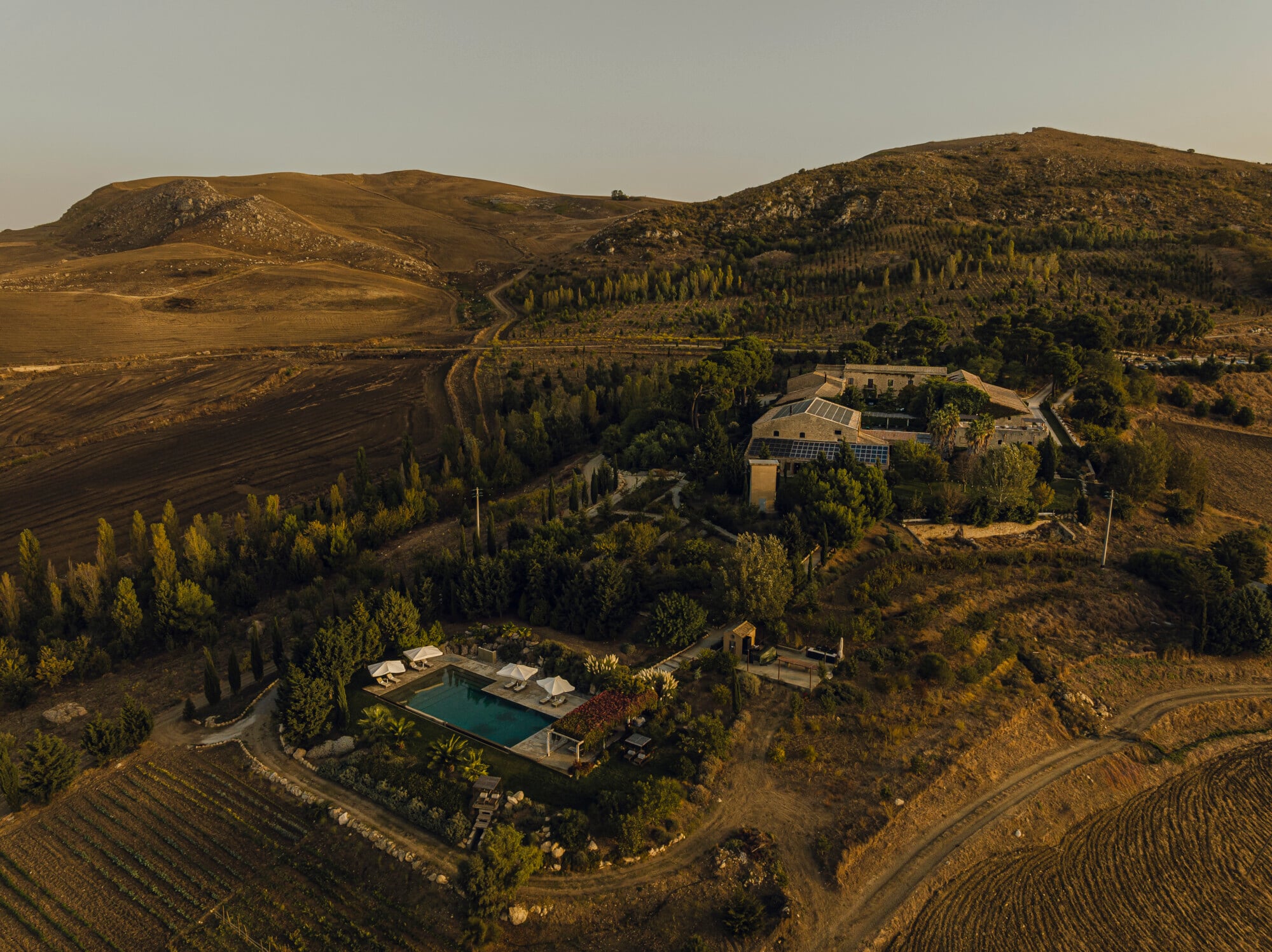 Susafa, Italy — Aerial view of modernist villa with solar panels nestled in forested Tuscan hills, infinity pool and terraces in golden evening light.