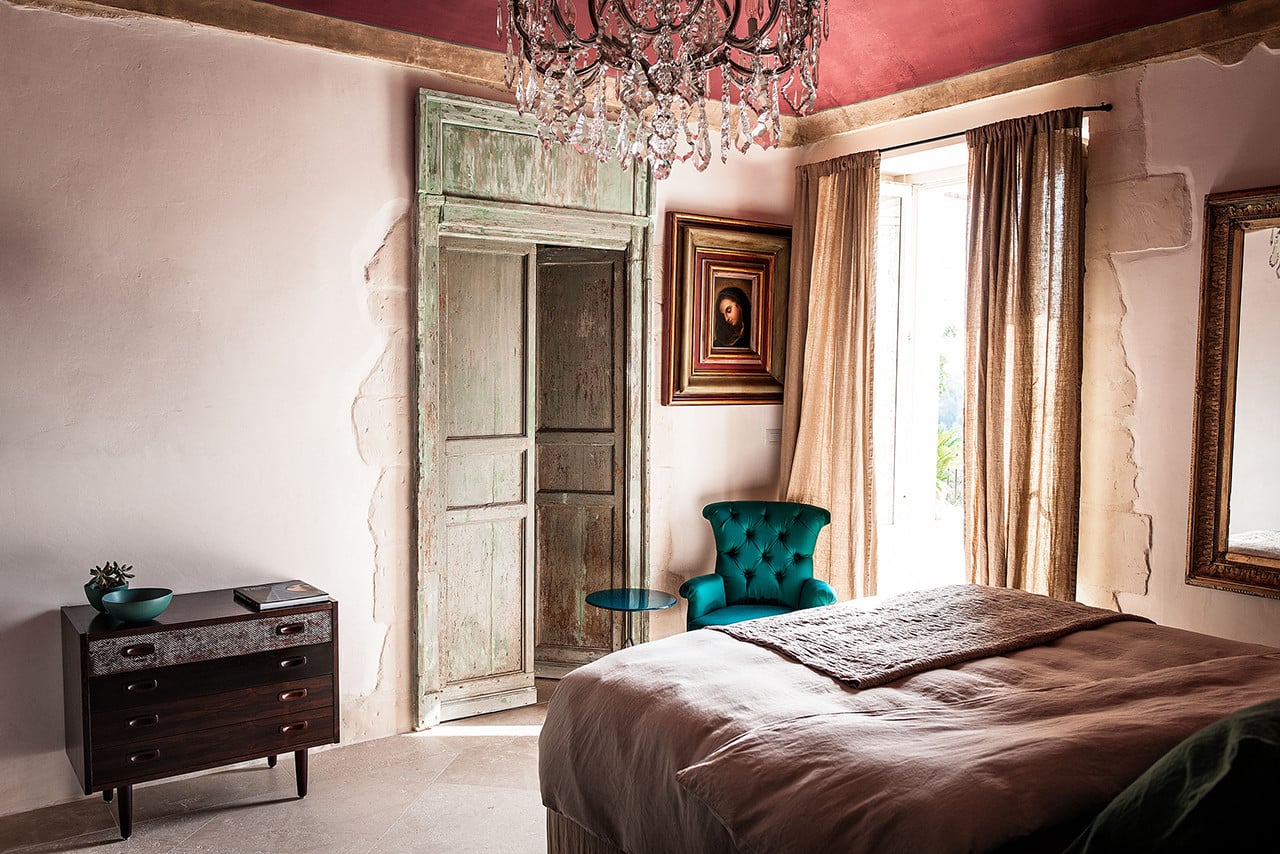 Dimora delle Balze, Europe — Bedroom with rose ceiling, distressed green doors, teal velvet chair, crystal chandelier, and warm natural light through linen curtains.