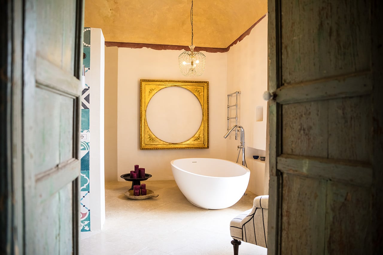 Dimora delle Balze, Europe — Minimalist bathroom with freestanding white oval tub, golden framed circular mirror, brass pendant light, terracotta walls, and dark wooden doors.