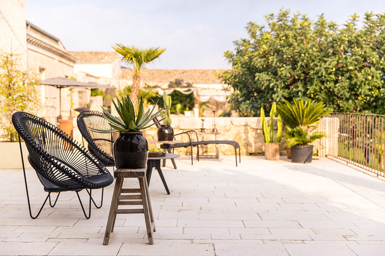 Dimora delle Balze, Europe — Black woven lounge chairs and potted succulents on pale stone terrace overlooking Tuscan villa architecture and cypress trees in golden afternoon light.