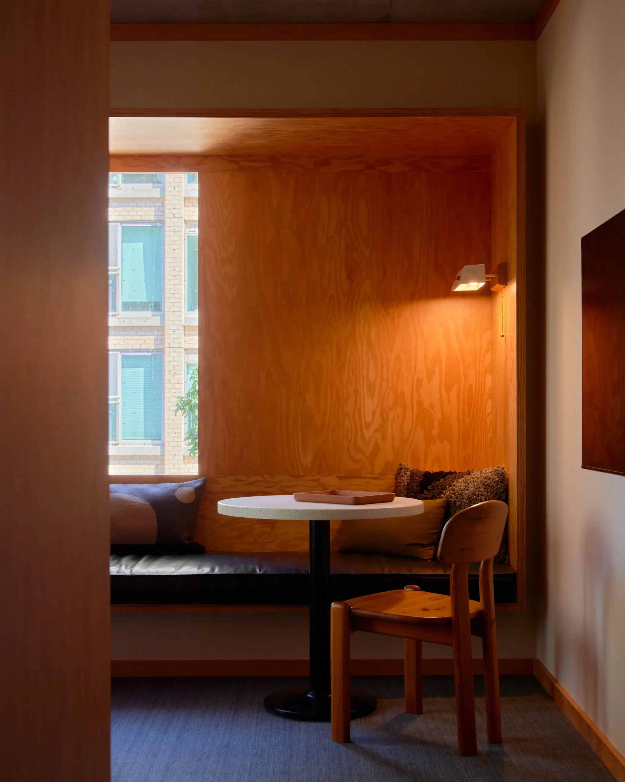 Ace Hotel Toronto, Canada — Warm wood alcove nook with built-in banquette, round table, mid-century chair, and natural light from city window.