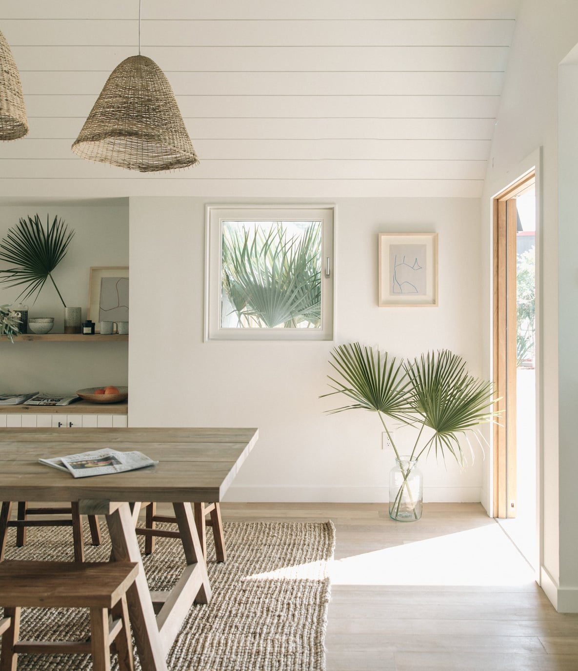 The Surfrider, North America — Bright dining room with woven pendant lights, natural wood table, potted palms, shiplap walls, and coastal modernist interiors flooded with daylight.