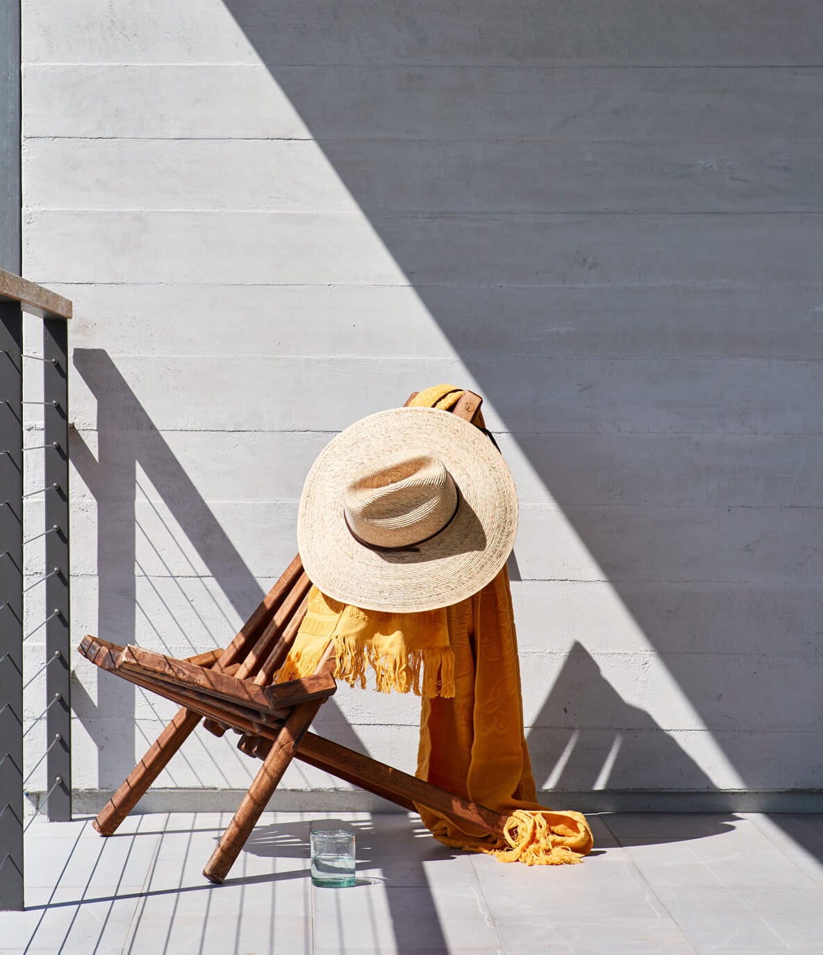 The Surfrider, North America — Straw hat and ochre linen draped over wooden chair against white brick wall, casting geometric shadows in bright sunlight, glass of water on terracotta tile floor.