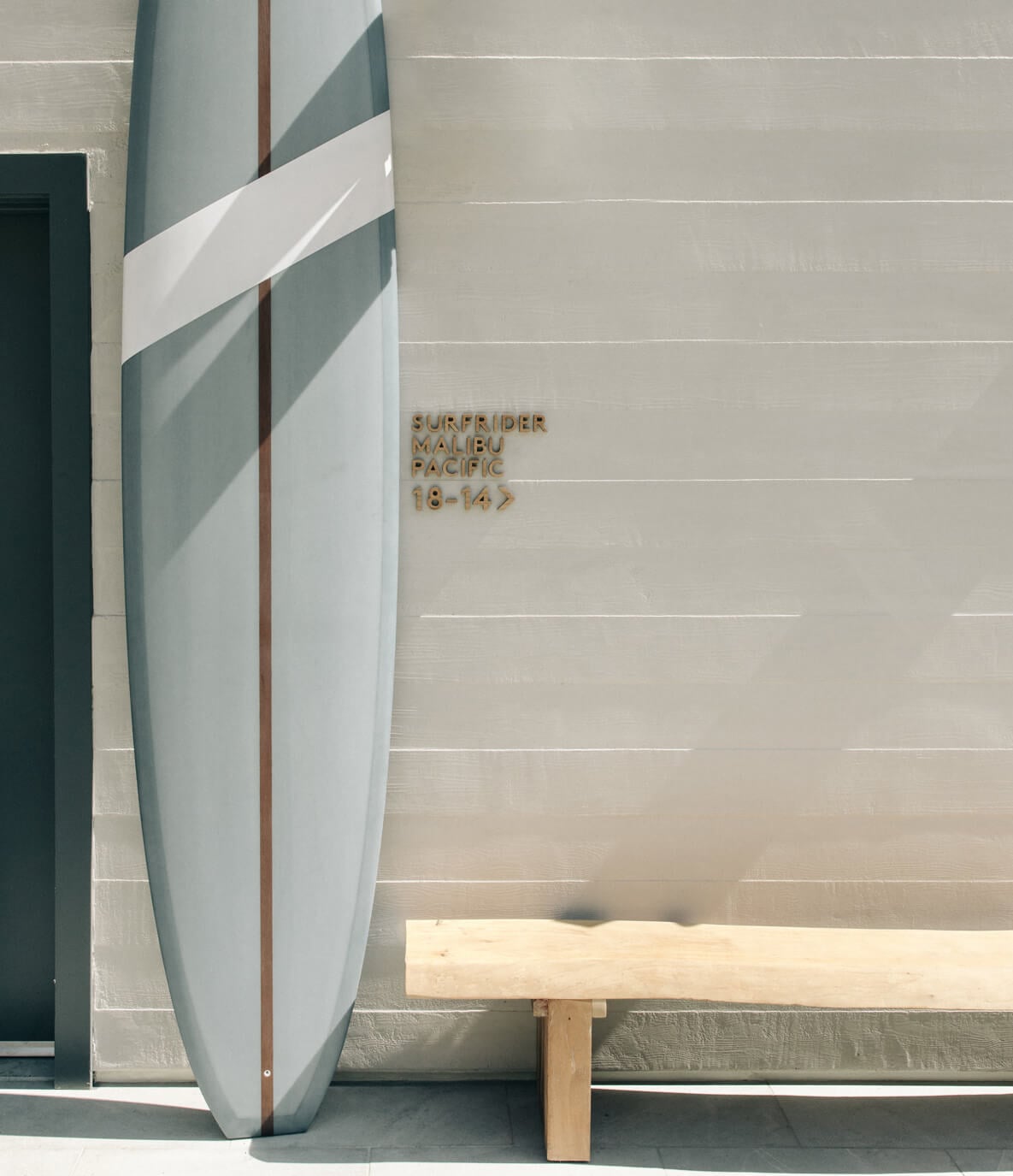 The Surfrider, Malibu, United States — Surfboard leaning against textured wall with striped flag and wooden bench in minimalist coastal interior.