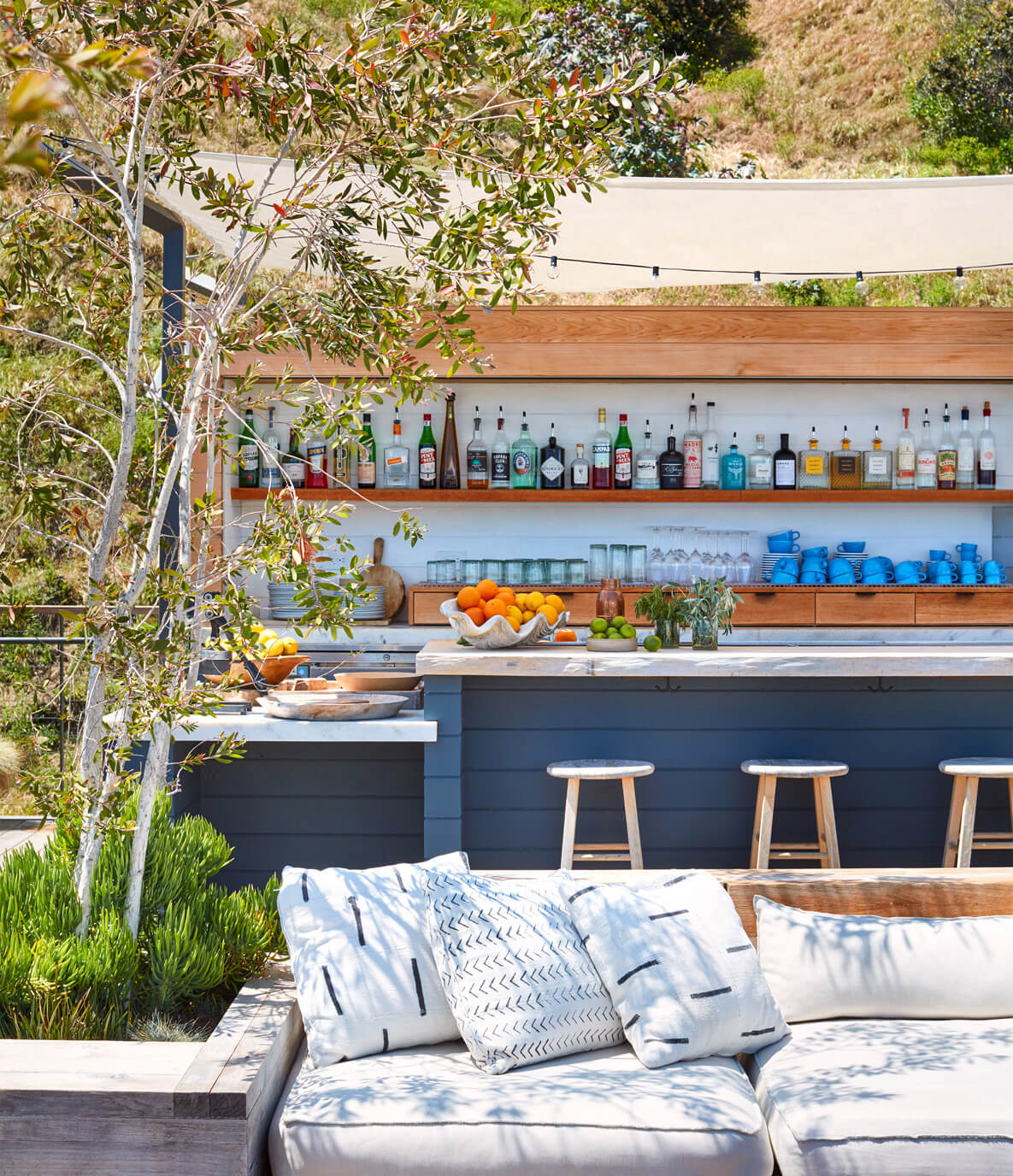 The Surfrider, North America — Outdoor bar with dark cabinetry, wood shelving, spirits bottles, blue glassware, citrus display, wooden stools, white cushioned seating, olive tree, hillside setting.