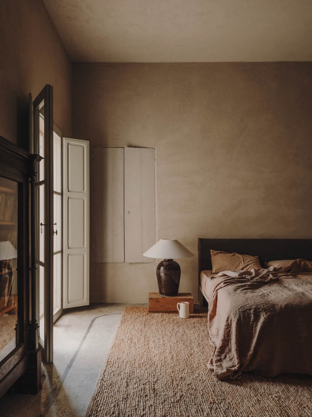 Casa Soleto, Europe — Minimalist bedroom with terracotta walls, natural linen rug, wooden nightstand, and cream bedding bathed in soft directional light.