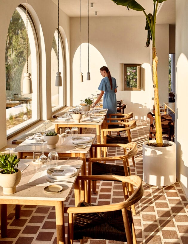 Parīlio Restaurant, Europe — Light-filled dining room with arched windows, wooden chairs, concrete tables, pendant lights, potted plants, and geometric floor pattern casting striped shadows.