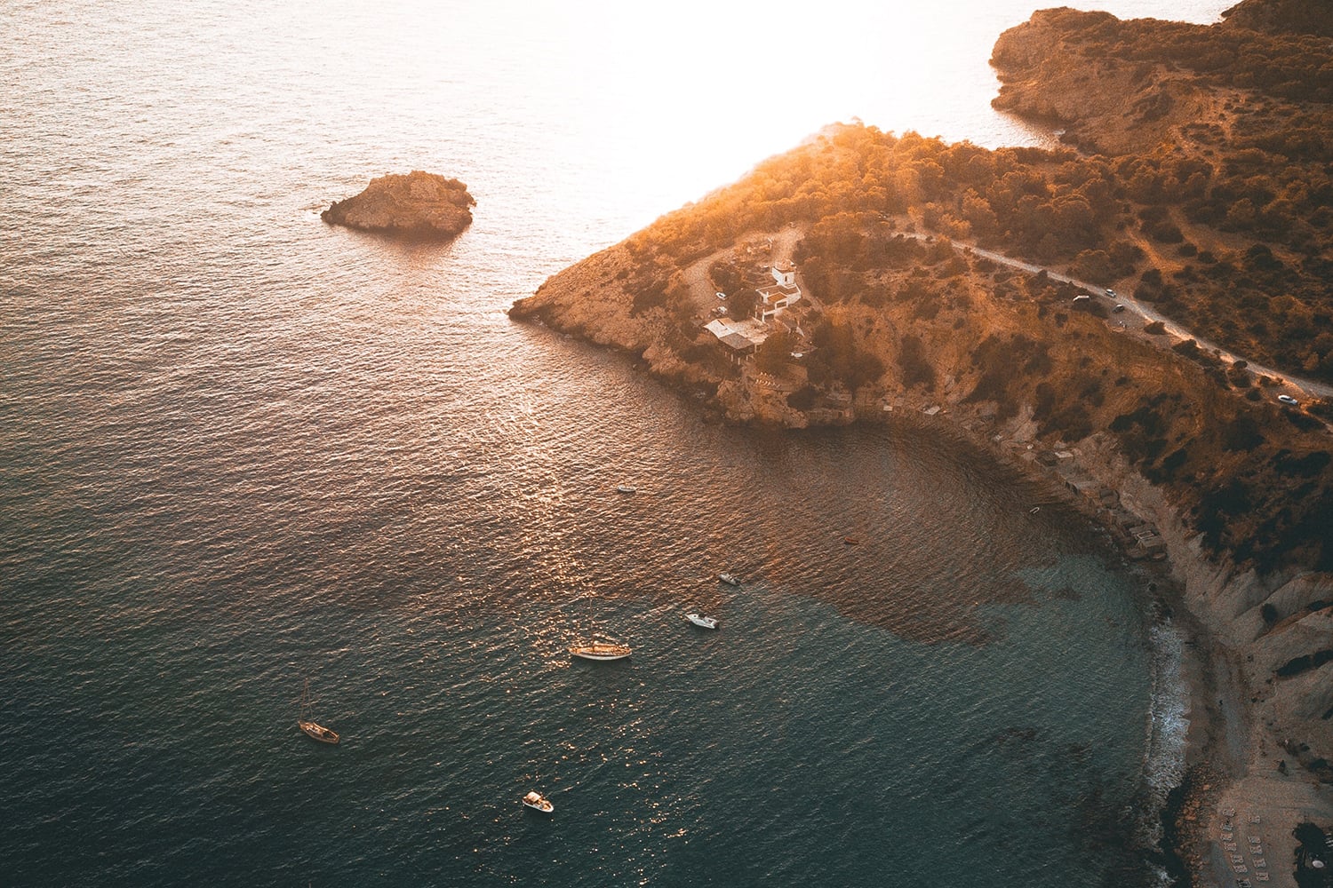 Six Senses: A New Dimension for Ibiza — Clifftop Mediterranean villa with terracotta stone overlooking golden-hour sea, moored boats, and rocky islets—luxury coastal architecture and landscape design.