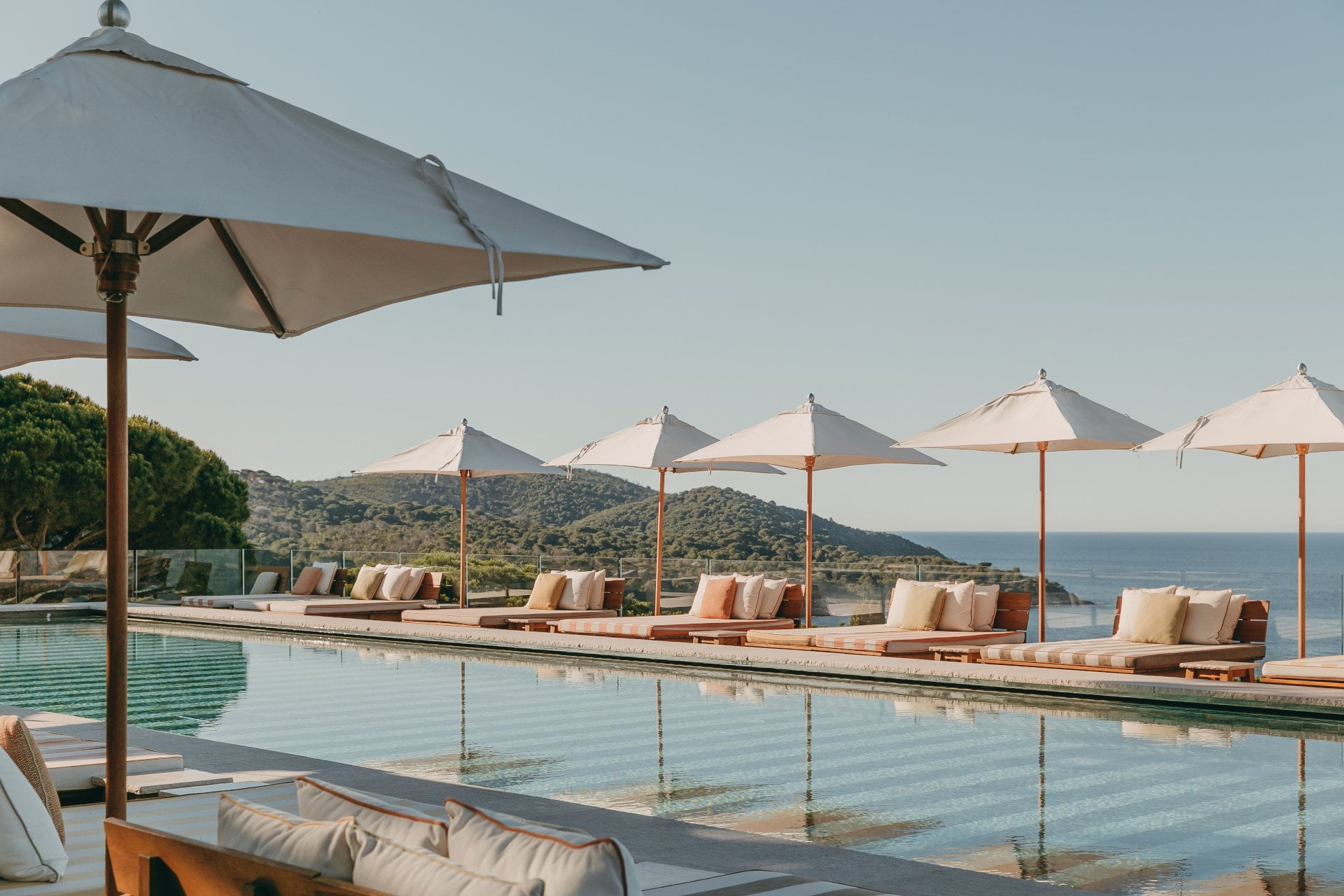 Lily of the Valley, Europe — Cantilevered poolside loungers beneath cream umbrellas overlooking terraced coastline and forested hills under clear sky.