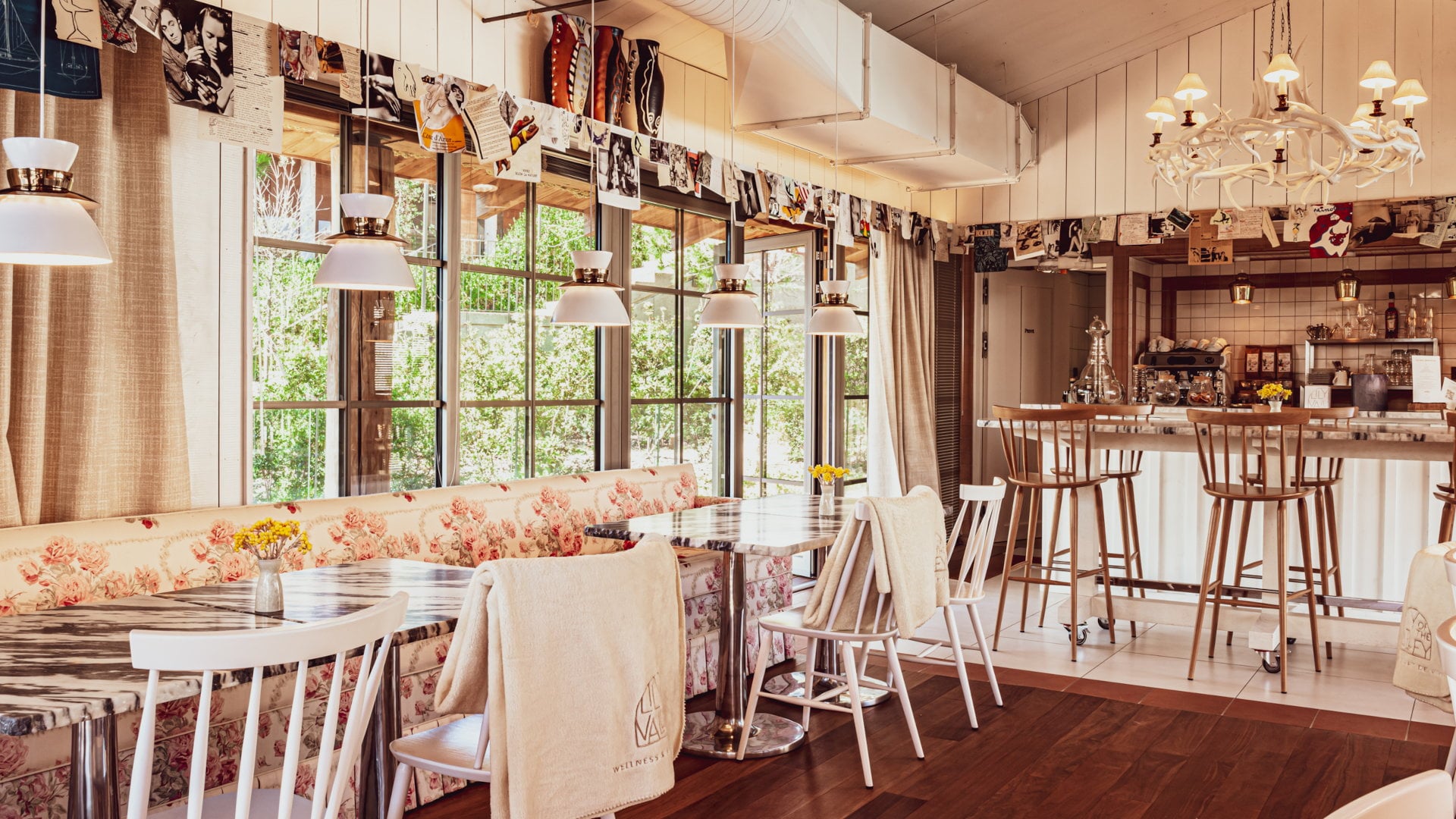 Club House, Europe — Bright dining room with pink floral banquettes, wooden chairs, pendant lights, large windows overlooking greenery, and bar counter with warm wood tones.