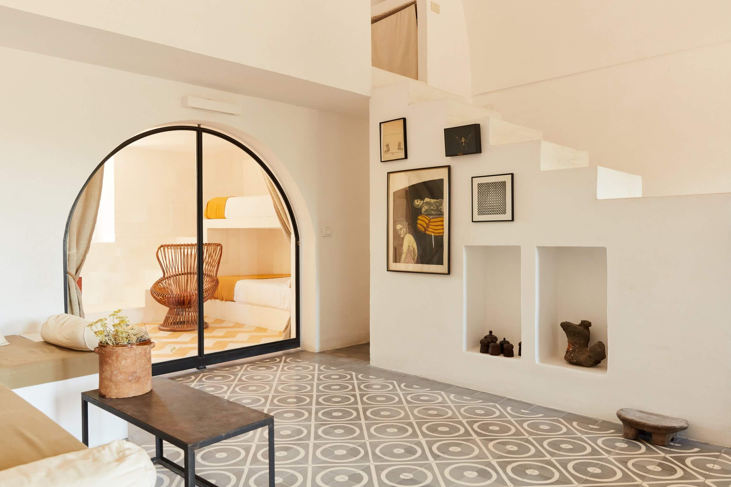 Parco Dei Sesi, Europe — Minimalist bedroom alcove with arched black-framed opening, woven chair, geometric floor tiles, floating shelves, and curated artwork gallery wall.