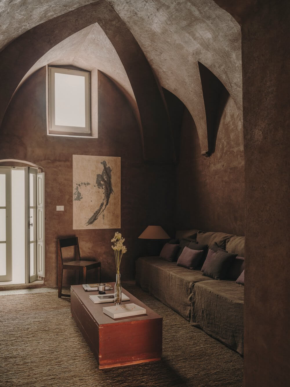 Casa Soleto, Europe — Terracotta interior with vaulted ceilings, arched window, linen sofa, wooden furnishings, and minimalist aesthetic.