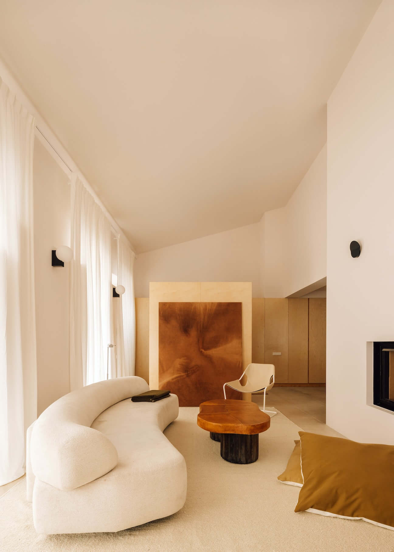 Casa Quatro, Europe — Minimalist living room with curved cream sofa, copper artwork, wooden table, sloped ceiling, and natural light from floor-to-ceiling windows.