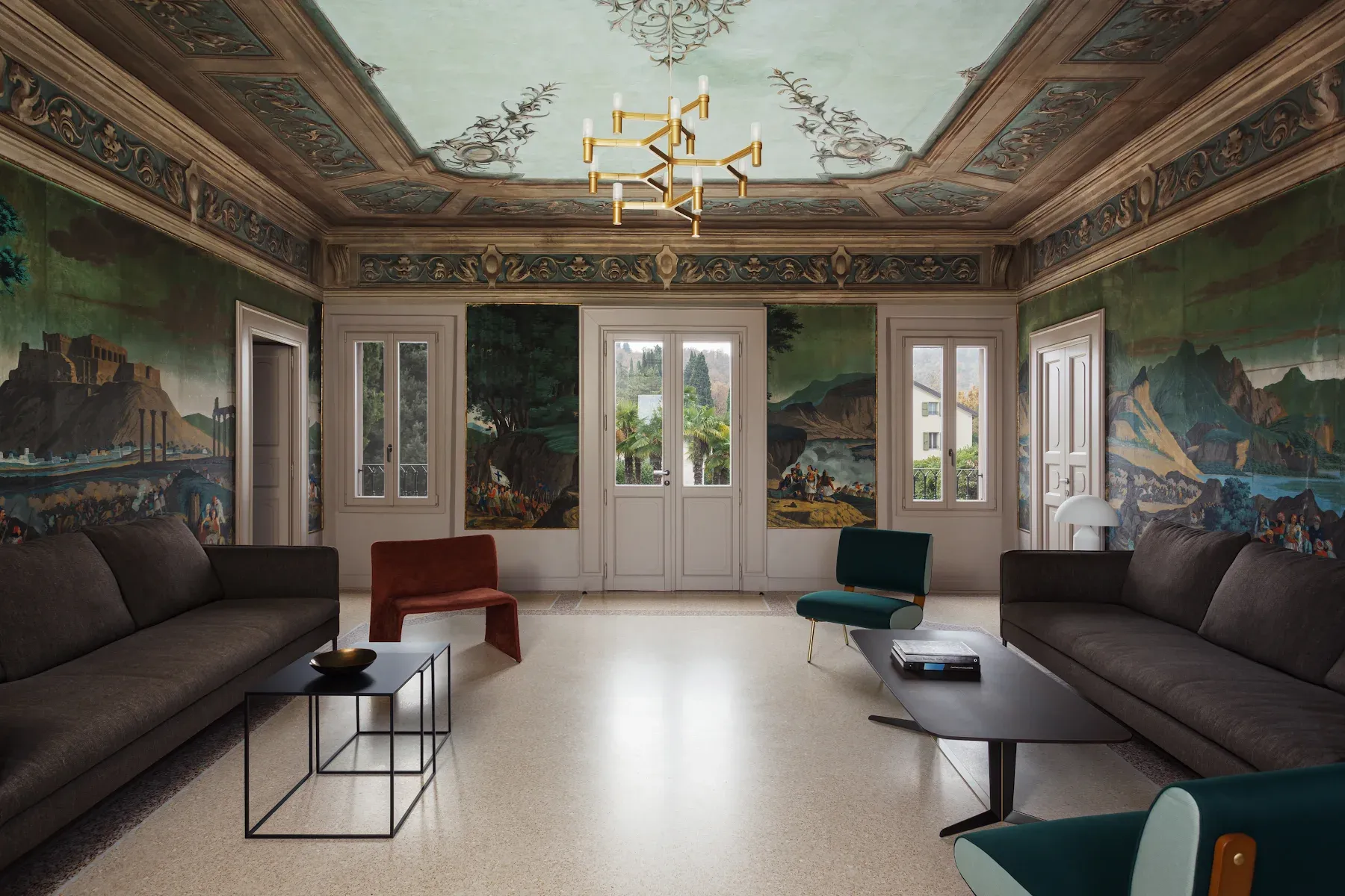 Roncolo 1888, Europe — Ornate coffered ceiling with frescoed panels, gallery-style salon featuring mountainous landscape murals, modern furnishings, gilt chandelier, natural light.