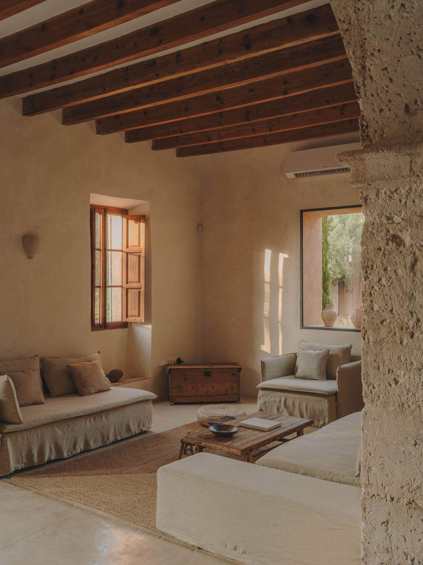 Can Lluïssó, Balearic Islands, Spain — Sunlit living room with exposed wooden beams, stone walls, linen seating, and terracotta windows overlooking Mediterranean courtyard.