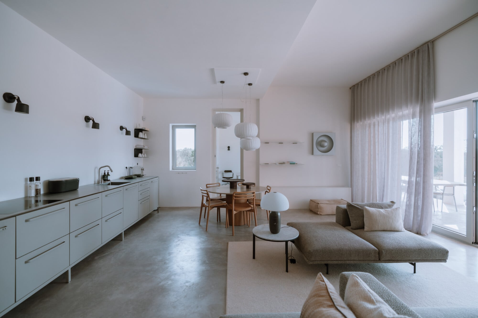Villa Quercia, Italy — Modern open-plan living space with white cabinetry, concrete floors, mid-century dining table, neutral linen seating, and floor-to-ceiling windows overlooking landscape.