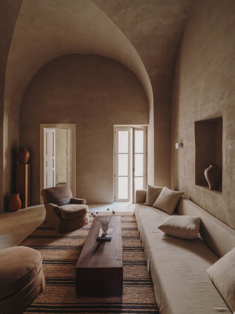 Casa Soleto, Europe — Minimalist living room with barrel-vaulted ceiling, linen seating, dark wood coffee table, terracotta vessels, and French doors opening to natural light.