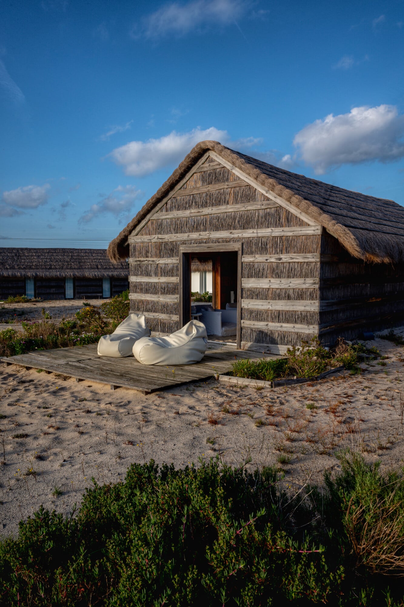 Casas na Areia, Europe — weathered timber beach pavilion with thatched roof, white bean bag seating on wooden deck, coastal dunes and blue sky.