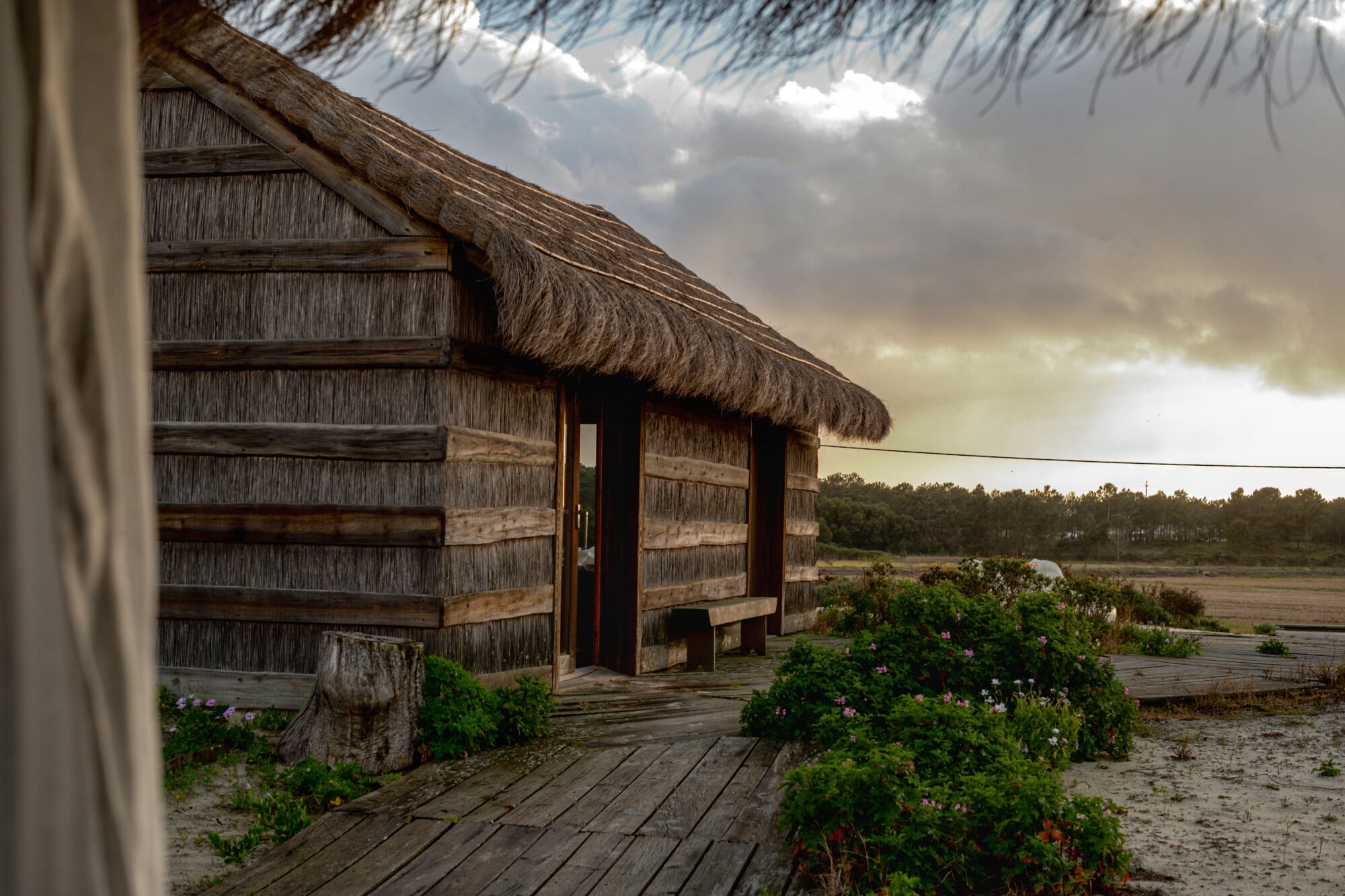 Casas na Areia, Europe — Thatched-roof wooden bungalow with weathered timber walls, sand courtyard, flowering plants, and lagoon views under dramatic sky.