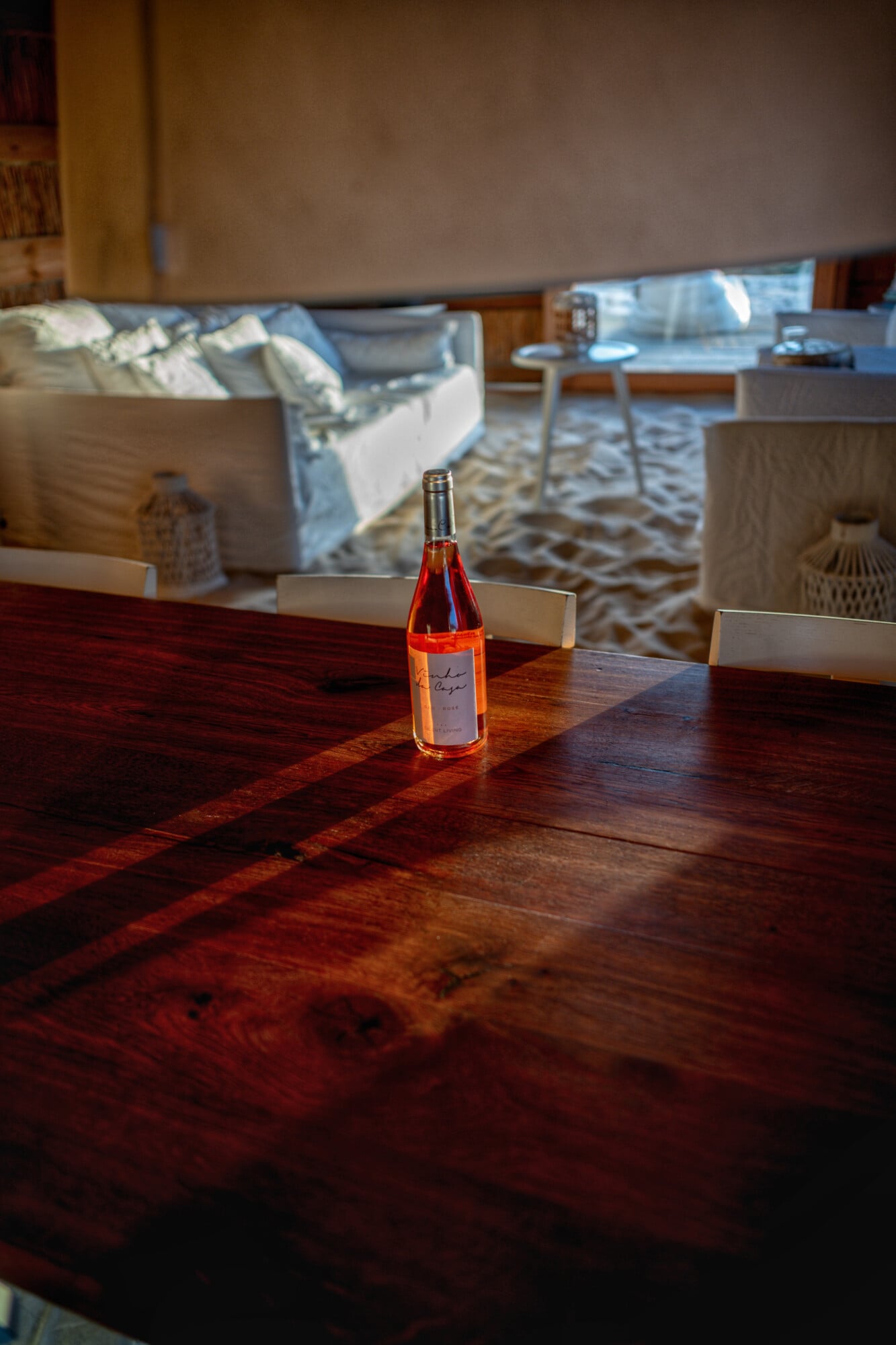 Casas na Areia, Europe — Rosé wine bottle on dark wood table with warm sunlight and modern living room interior in soft focus.