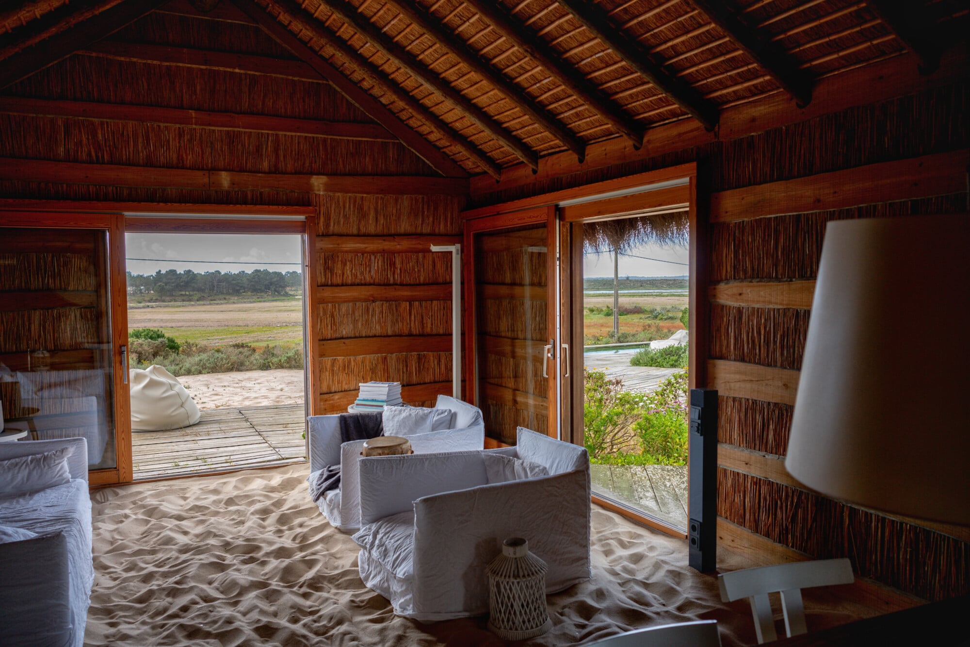 Casas na Areia, Europe — Sandy interior with rattan walls, white linen seating, wooden beam ceiling, and views across wetlands through open doors.